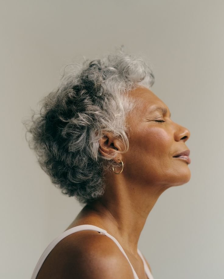 Profile of a joyful older woman with silver, curly hair, wearing small hoop earrings and a white tank top, smiling with her eyes closed.