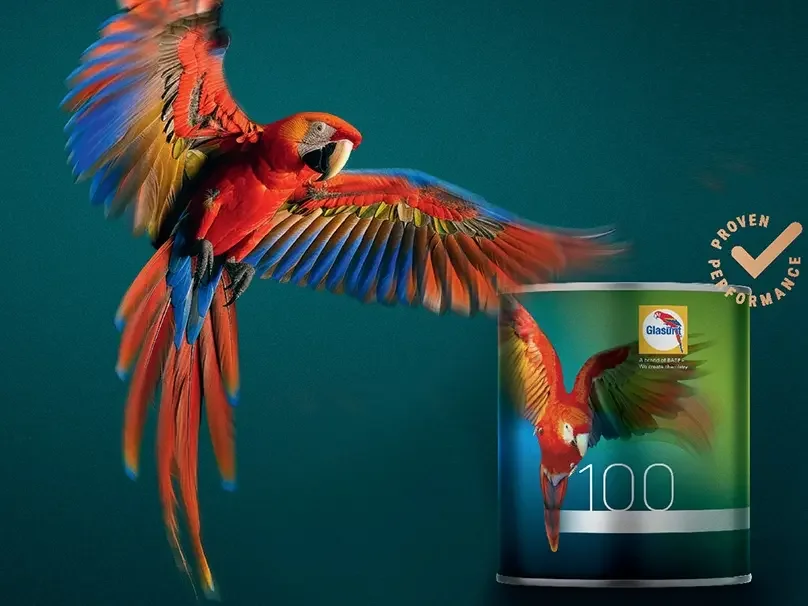 A colorful parrot with red, blue, yellow, and green feathers flying next to a can of Glasurit 100 paint, which features a similar parrot design on its label.