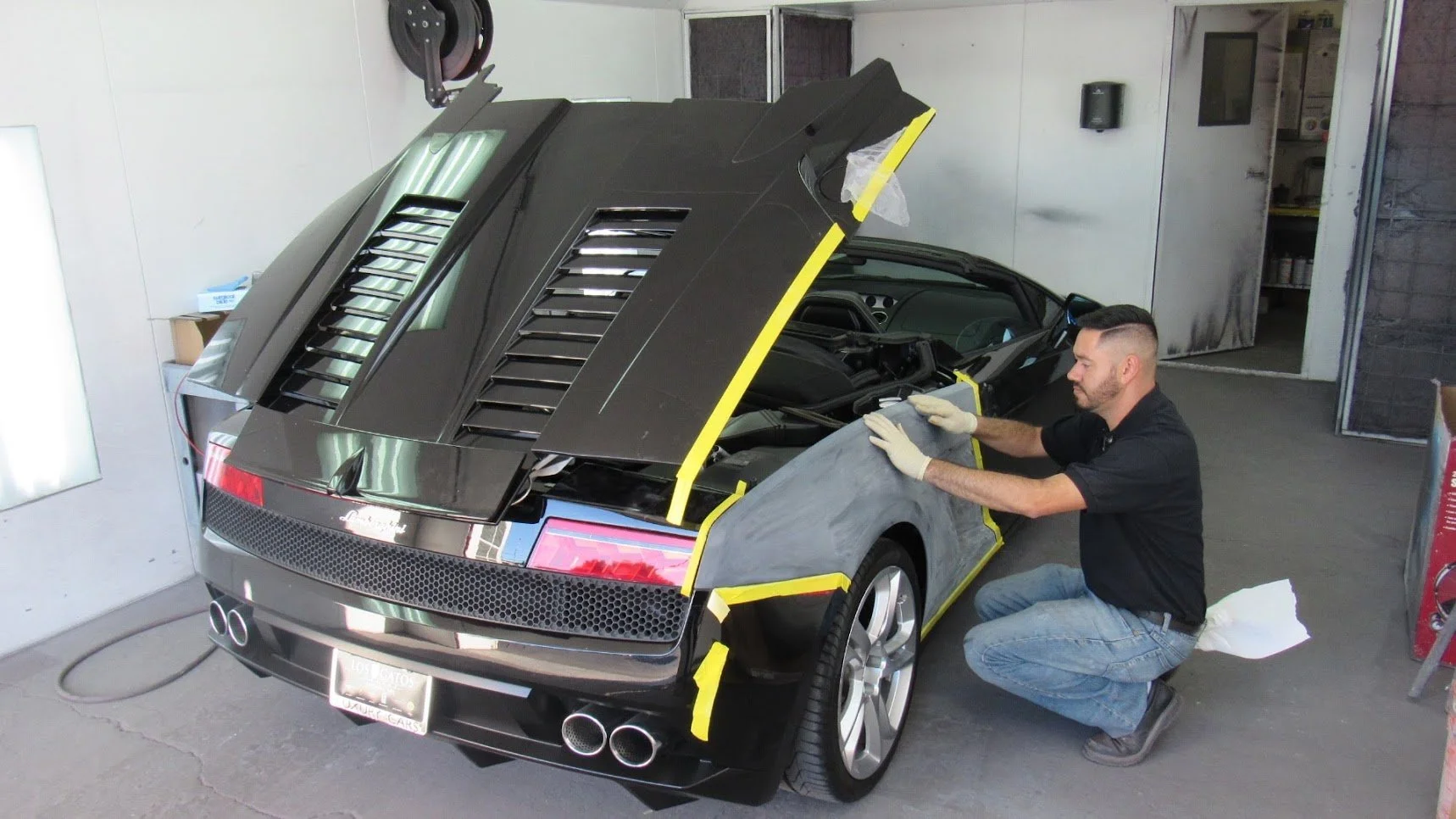 A man working on a black sports car inside a garage, installing or repairing parts with masking tape and protective gloves.
