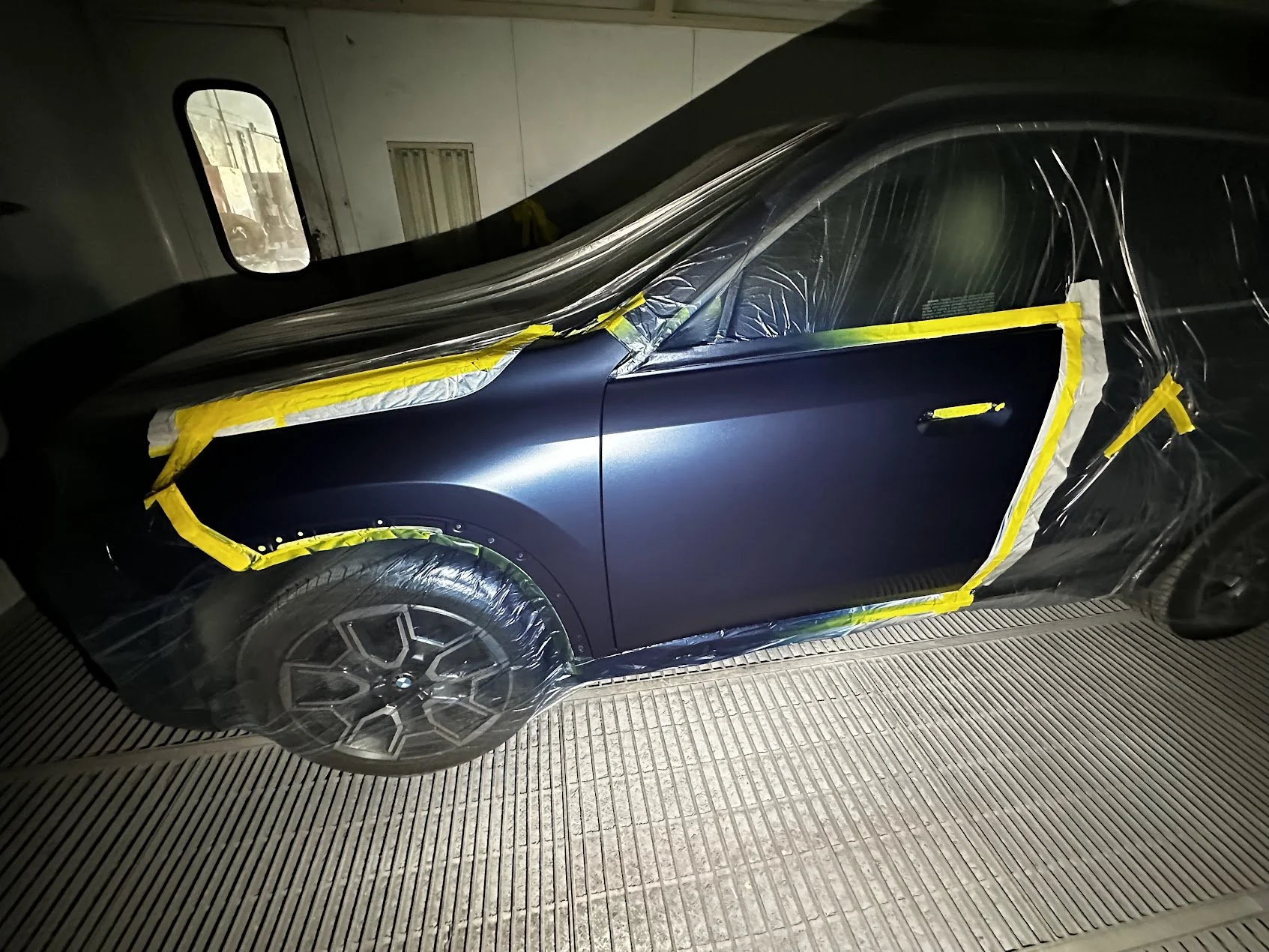 A dark blue BMW car is covered with protective plastic and masking tape, likely in a workshop or paint booth, preparing for painting or detailing work.