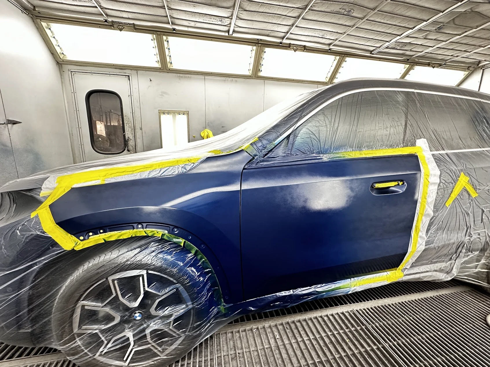 A blue car is inside a paint booth, covered with plastic and masking tape, likely being prepared for painting or touch-up.