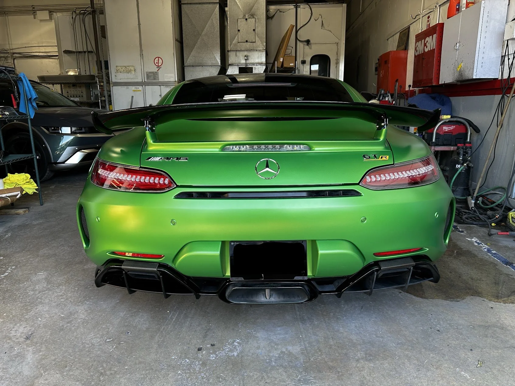 A bright green Mercedes-AMG GT Black Series sports car with a large rear wing is parked inside a garage, with another black and gray sports car to its left. The garage contains various tools, equipment, and storage cabinets on the walls.