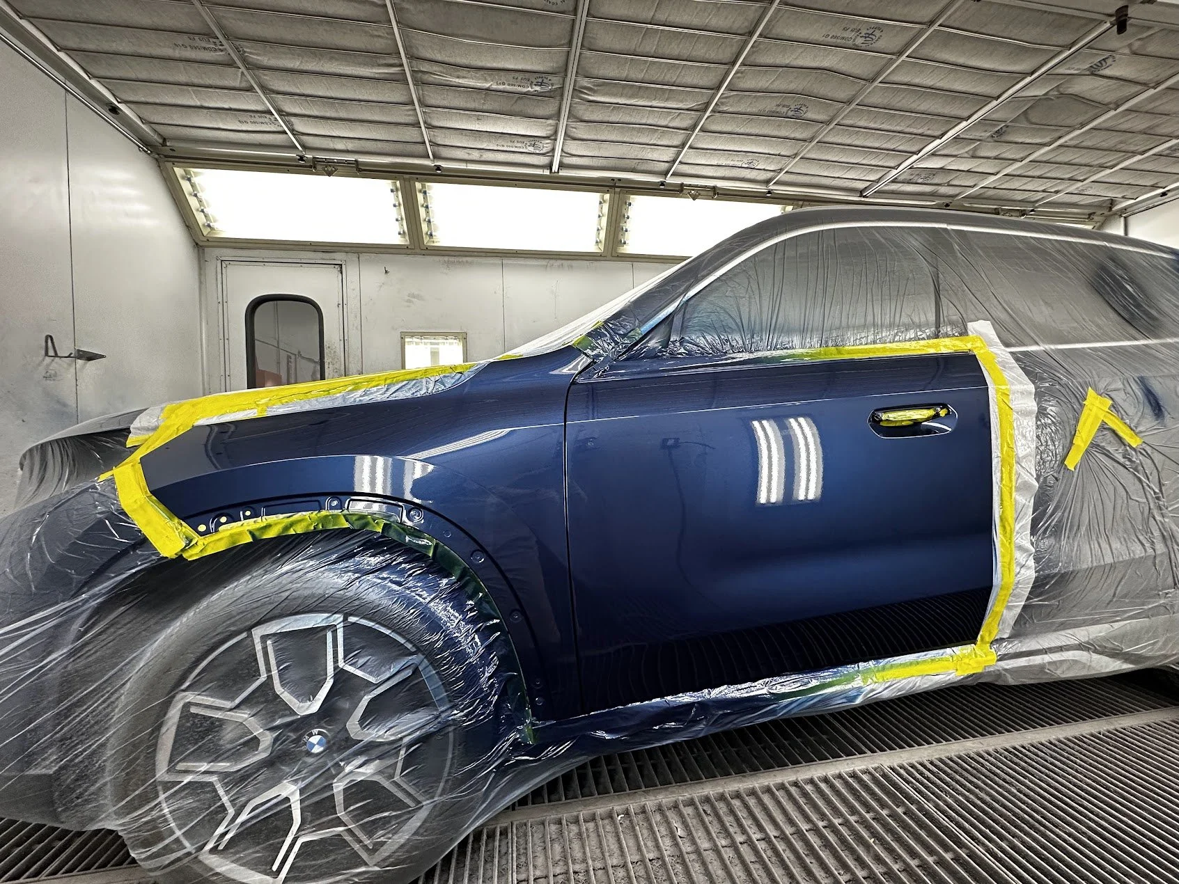 A blue car, likely a sports model, is inside a paint booth, covered with plastic and yellow masking tape, indicating it is in the process of being painted or detailed.