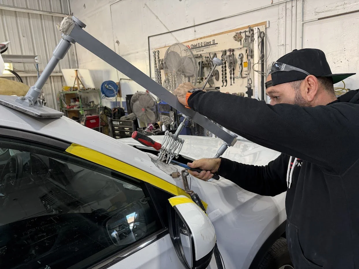 A man wearing safety glasses and a baseball cap working on a white car in a garage or workshop. He is using tools on the car's roof, which is covered with clamps and a metal frame for testing or repairs. The background shows shelves with tools and equipment, and a pegboard with various hanging tools.