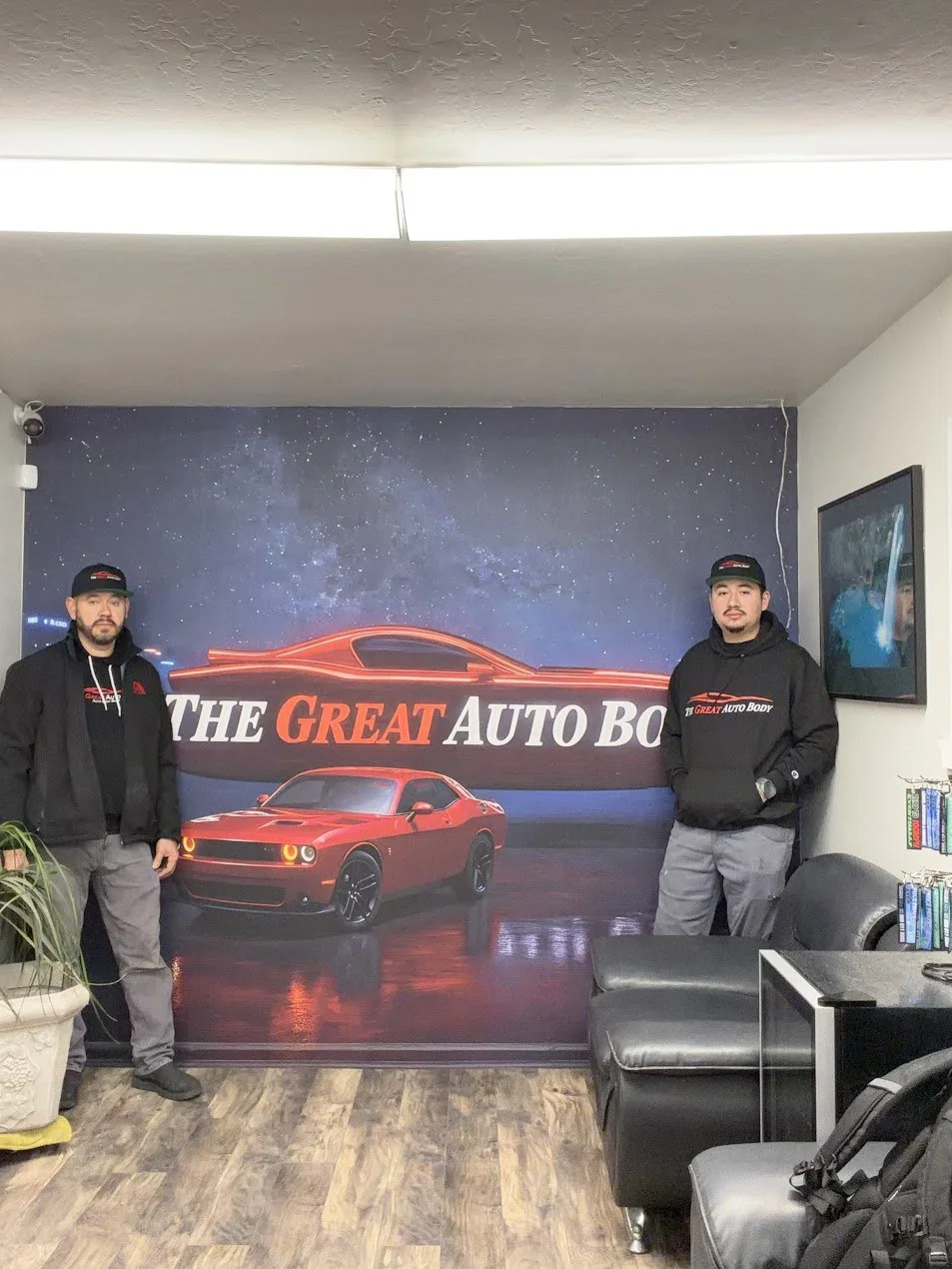 Two men standing in front of a large banner with a red sports car and the words "The Great Auto Body" in a room with wood flooring and a television on the wall.