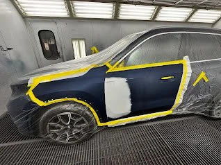 A dark blue car in a paint booth, with parts masked off in yellow tape and primer applied to the door.