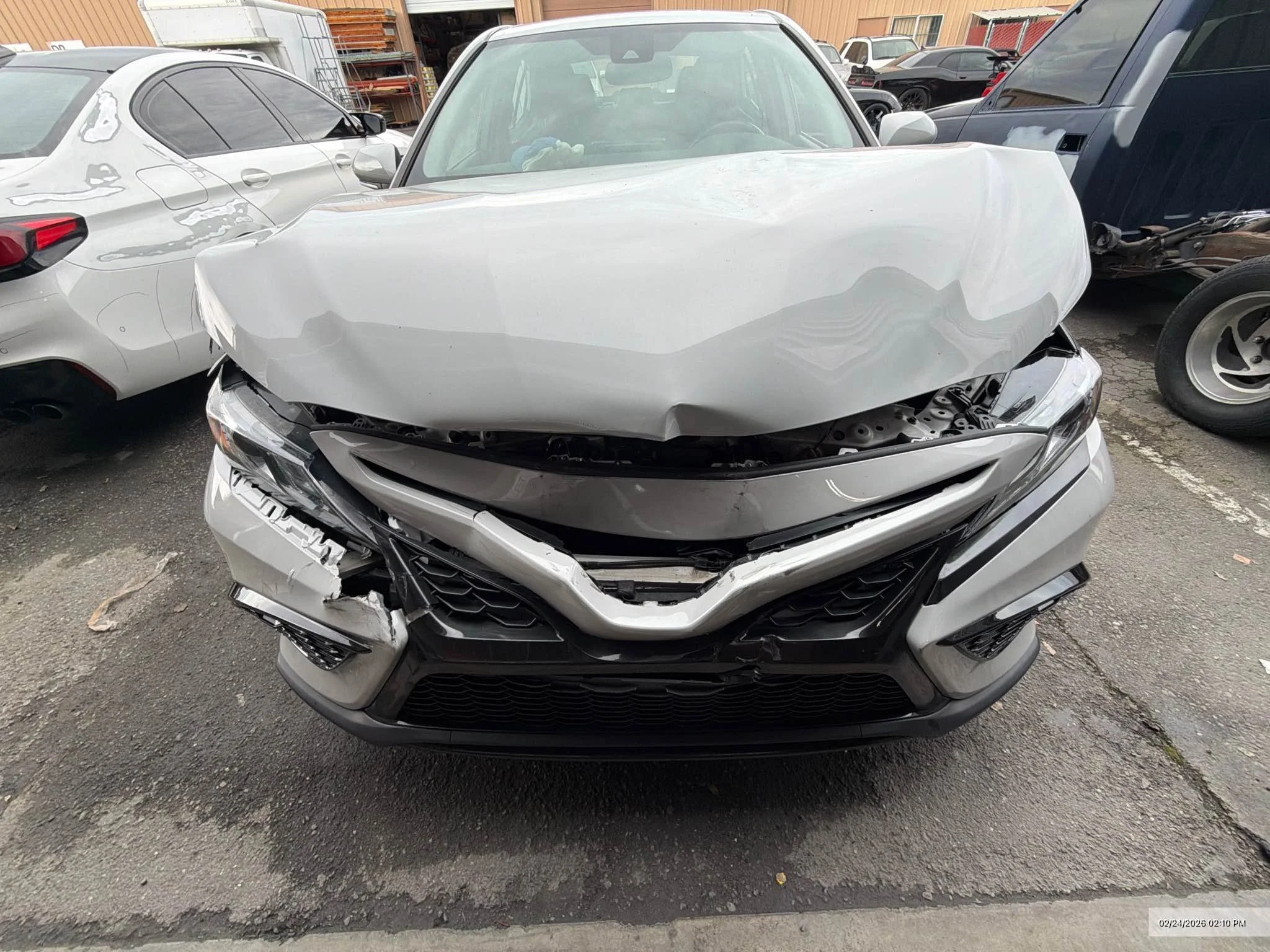 Front view of a gray car with front-end damage, missing bumper, in an outdoor lot surrounded by other vehicles and a storage building.