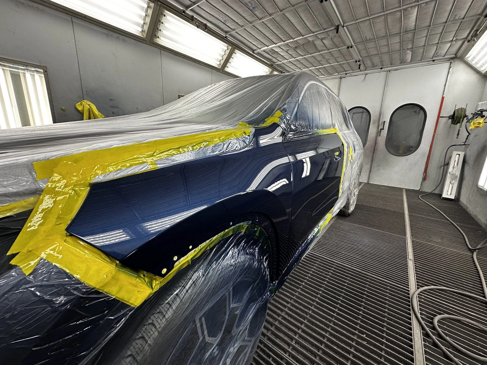 A black car in a paint booth, covered with masking tape and plastic sheeting, ready for painting.