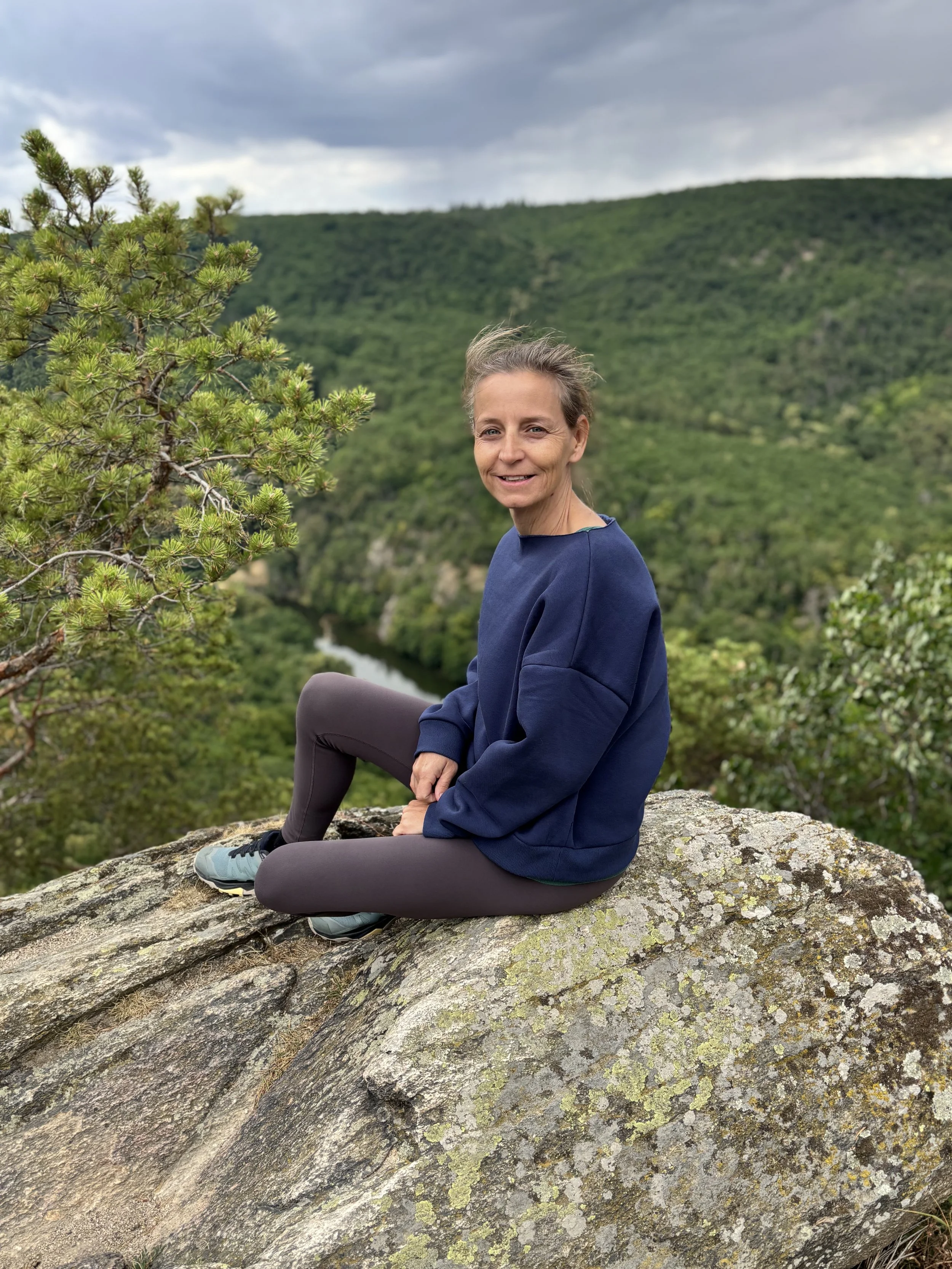 A woman sitting on a large rock outdoors with a lush green hillside and a river in the background, wearing a blue sweatshirt and brown pants.