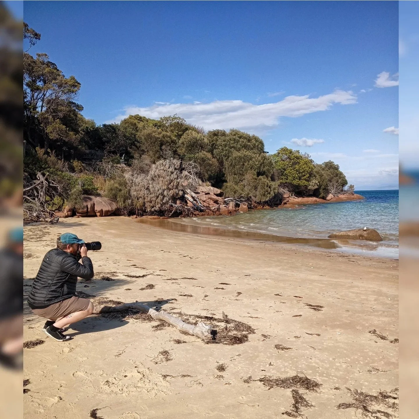 A person kneeling on a sandy beach taking a photo with a camera, with trees and rocks along the shoreline and a blue sky with some clouds.