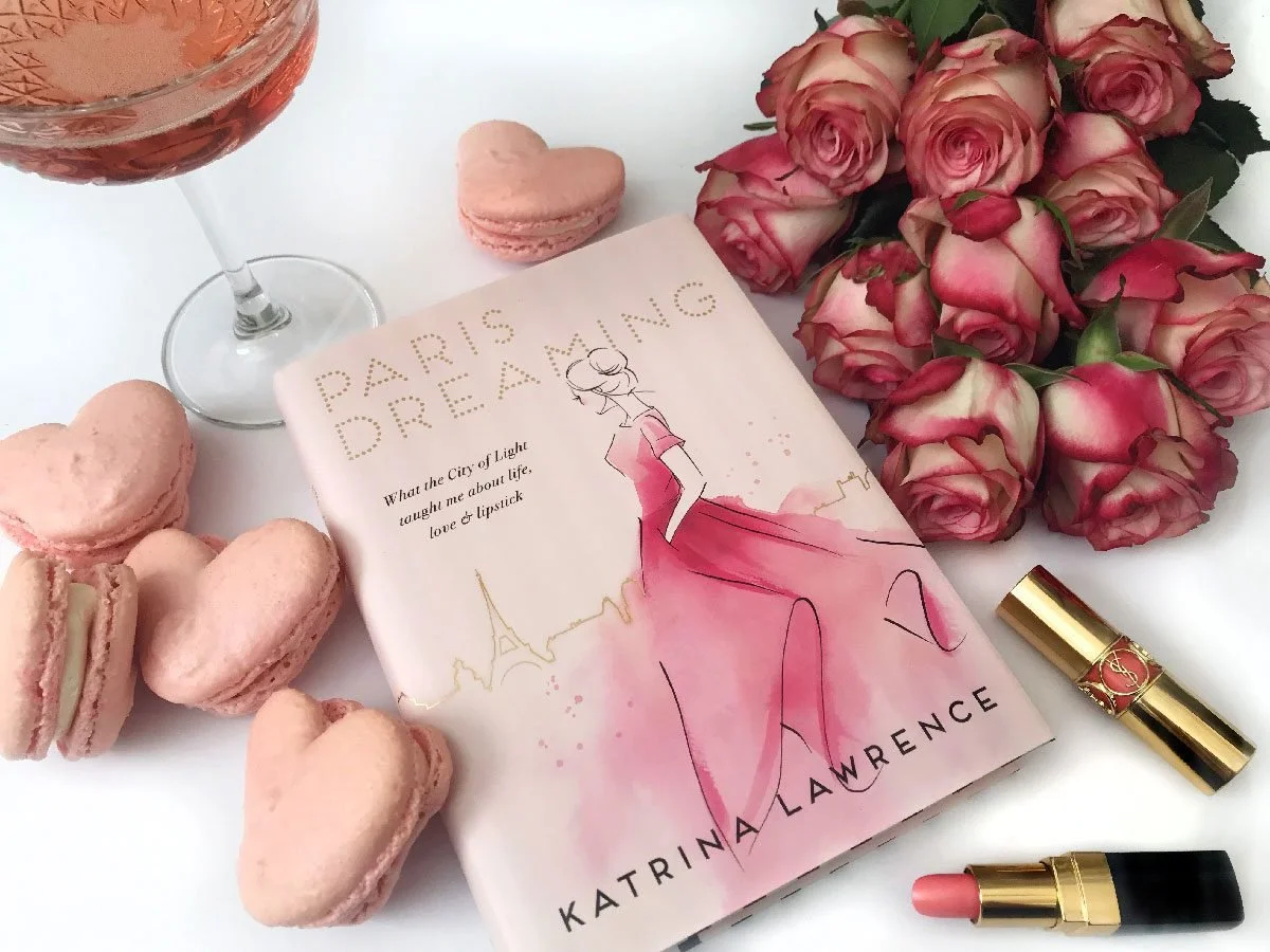 A flat lay of a romantic scene with pink roses, heart-shaped macarons, a glass of rosé wine, a book titled "Paris Dreaming" by Katrina Lawrence, a gold and red lipstick, and a pink lipstick, set against a white background.