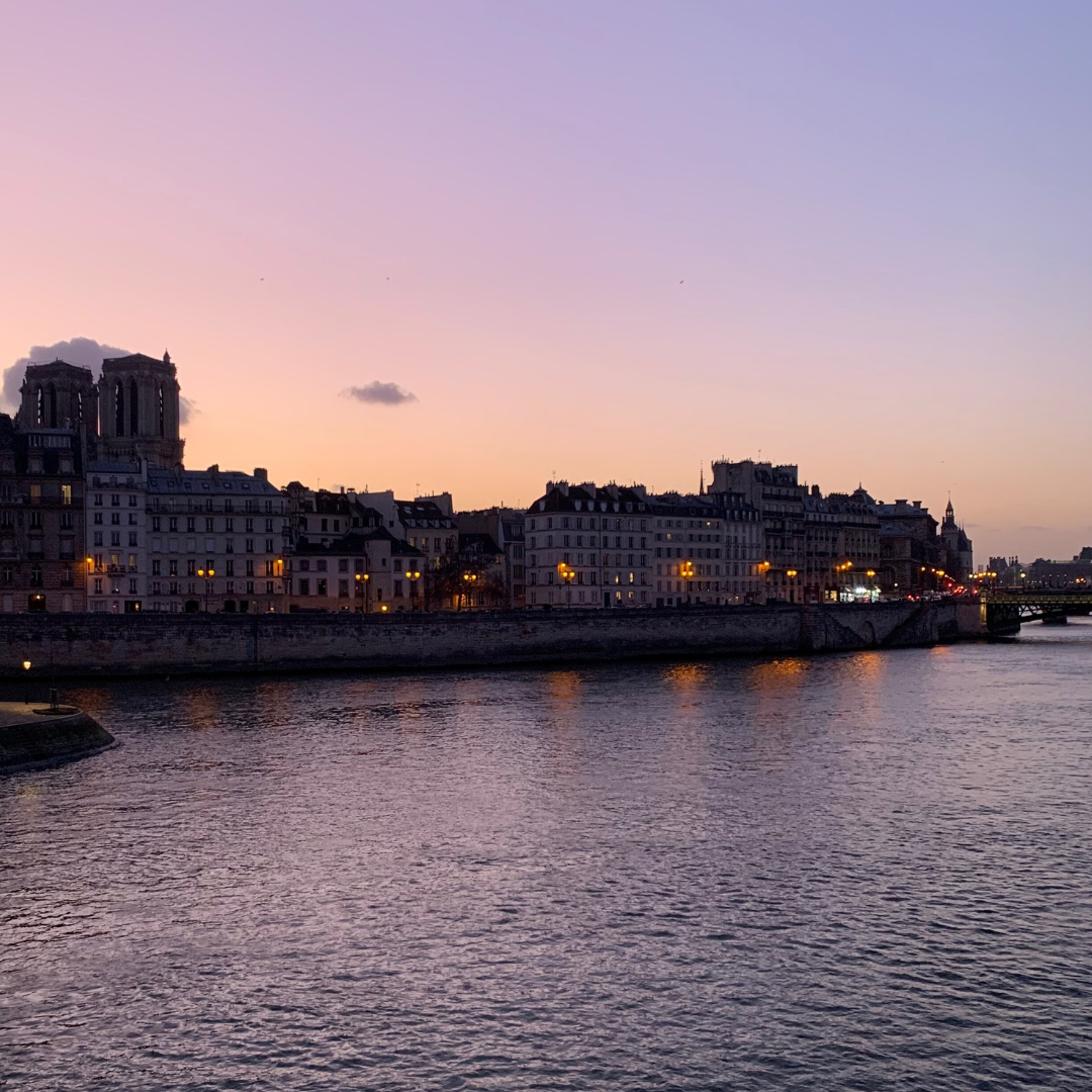 Sunset over the Seine River in Paris, with historic buildings and Notre Dame Cathedral in the background. The sky has a pink and purple hue, and city lights reflect on the water.