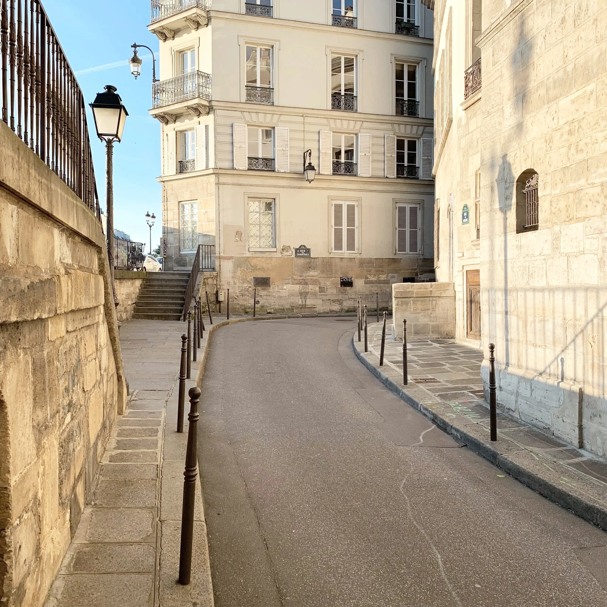 A narrow European street with stone stairs, stone buildings with balconies, street lamps, and bollards on the sidewalk.