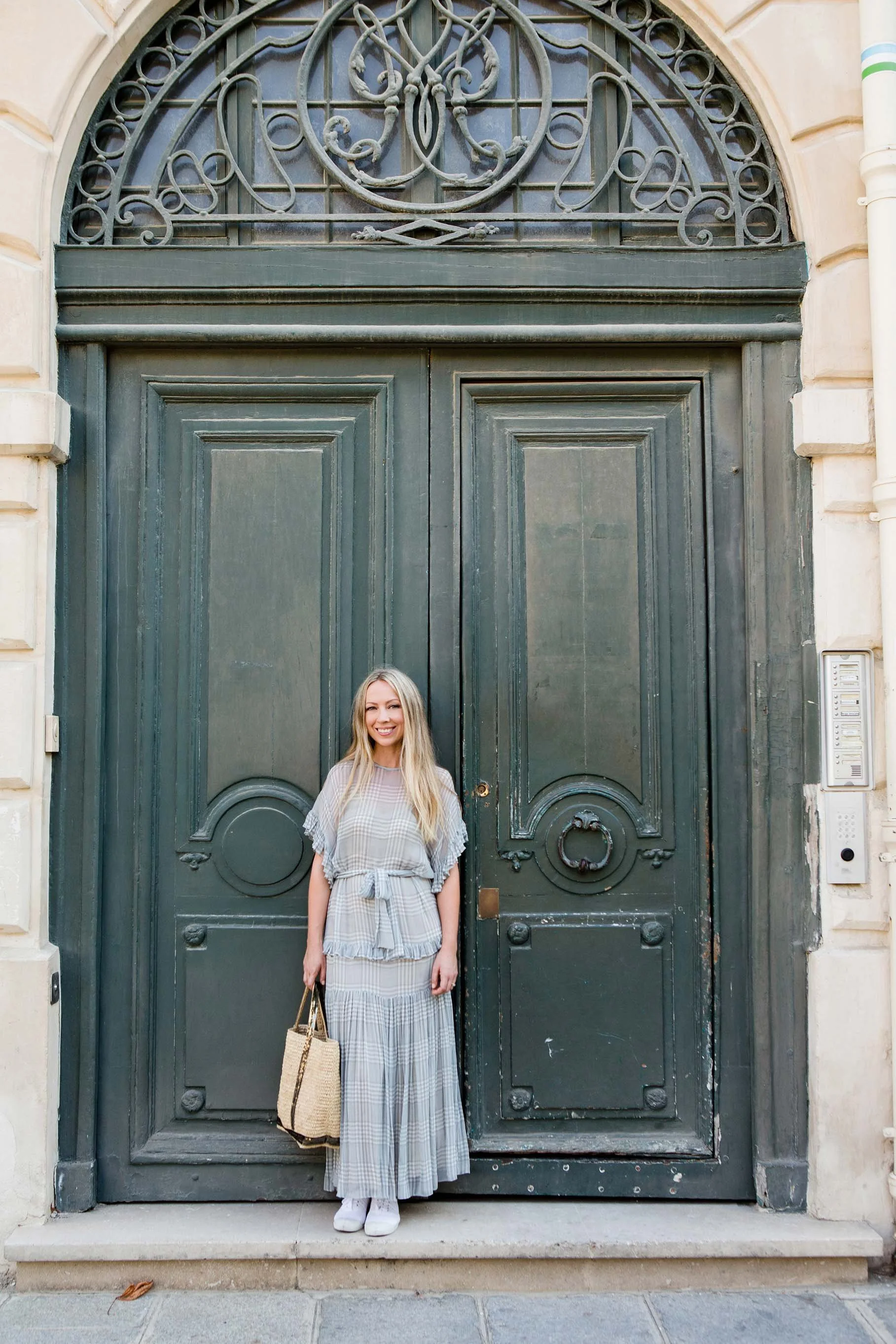 A woman with blonde hair wearing a plaid dress and white sneakers standing in front of a large ornate black door with intricate metalwork and a crest at the top, holding a woven bag and smiling.