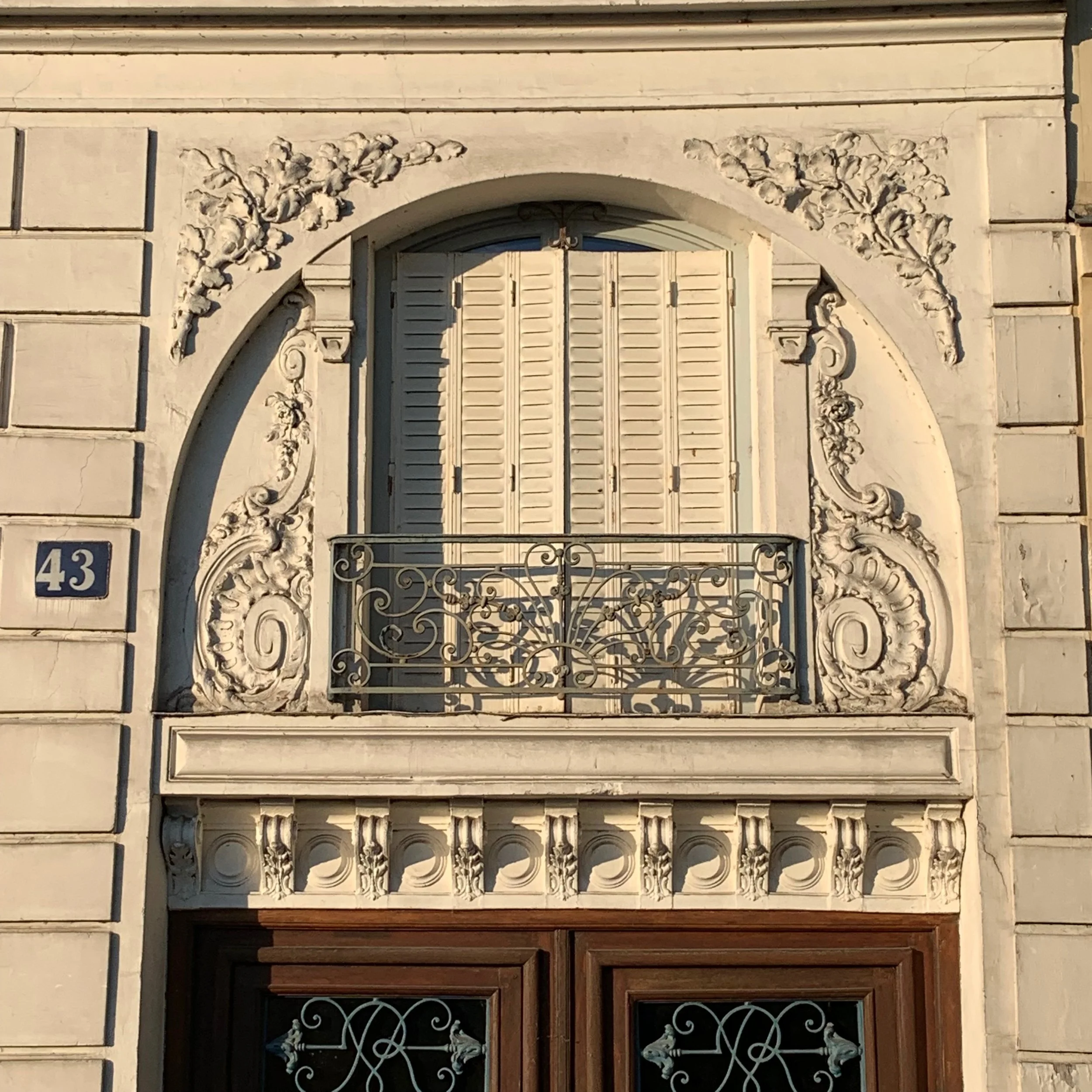 An ornate building facade with a rounded arch window or door, decorative floral and scroll carvings, and a wrought iron balcony or grille, with the number 43 on a blue plaque.