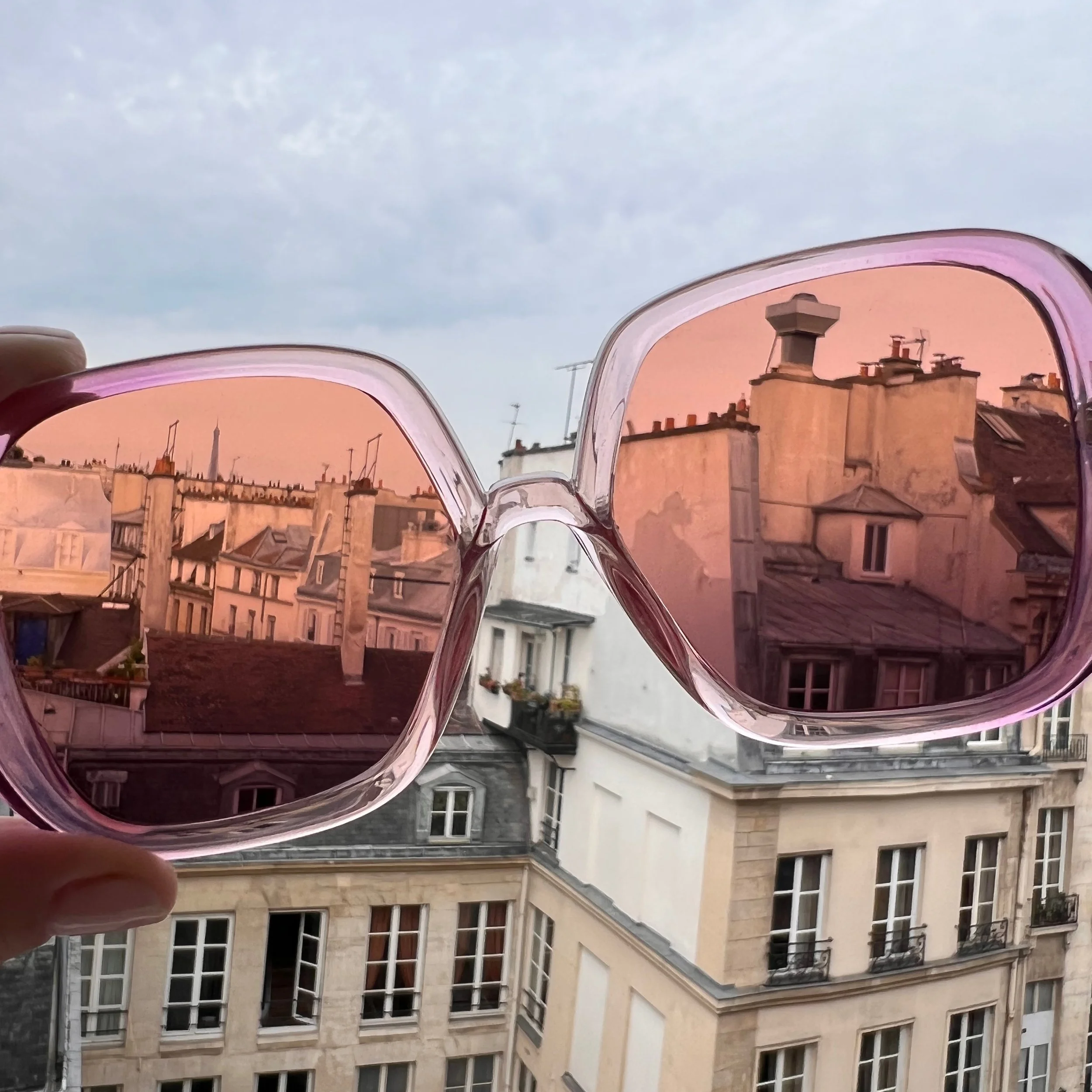 Reflection of a cityscape with buildings and sky in pink-tinted sunglasses held in a person's hand.