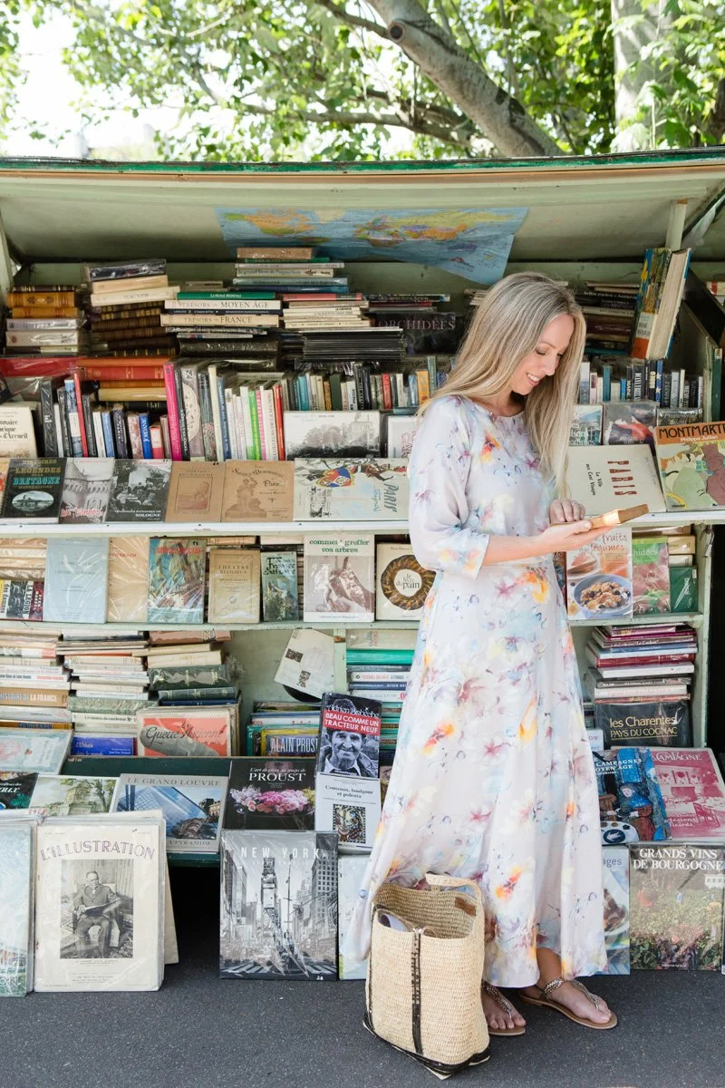 A woman in a light, floral dress stands outside a book stand, looking at her phone, with a basket at her feet, and various books stacked on shelves behind her.