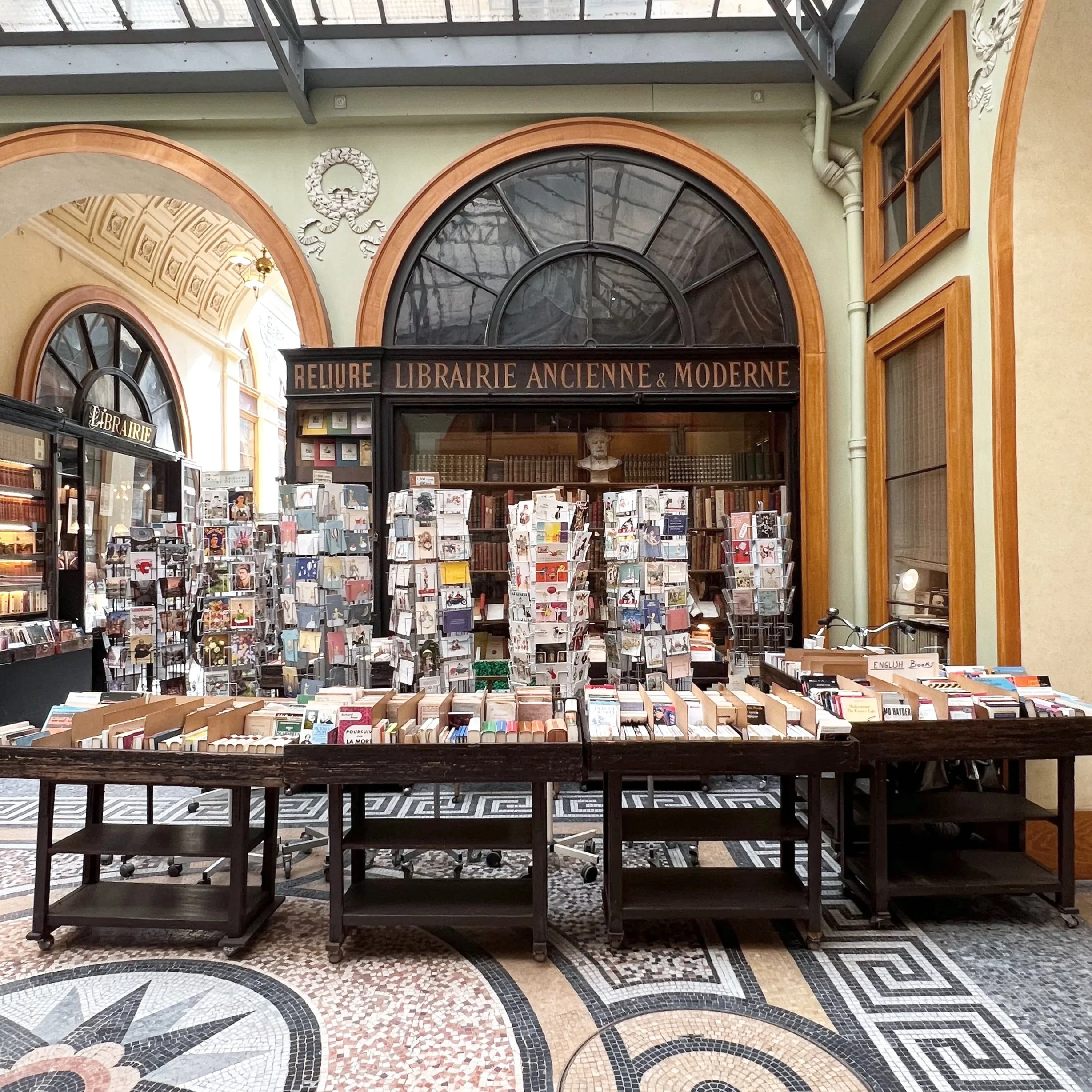 Interior of a vintage bookstore with a display table of books and an arched window above. The sign reads 'Relure Librairie Ancienne & Moderne,' and the floor has a mosaic pattern.