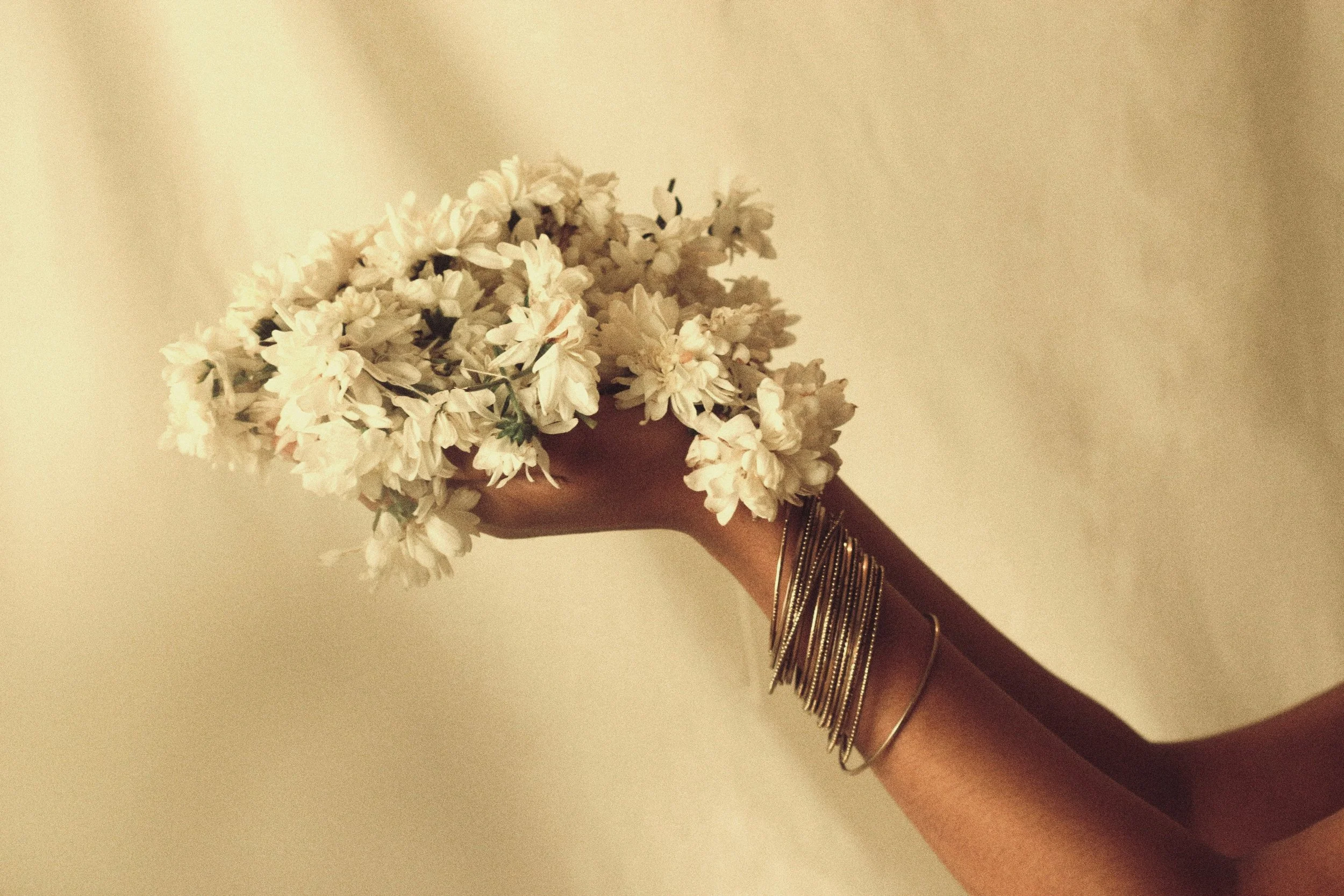 A person with Bangladeshi skin tone is holding a bouquet of white flowers, wearing multiple silver bangles on their wrist, against a plain beige wall background.