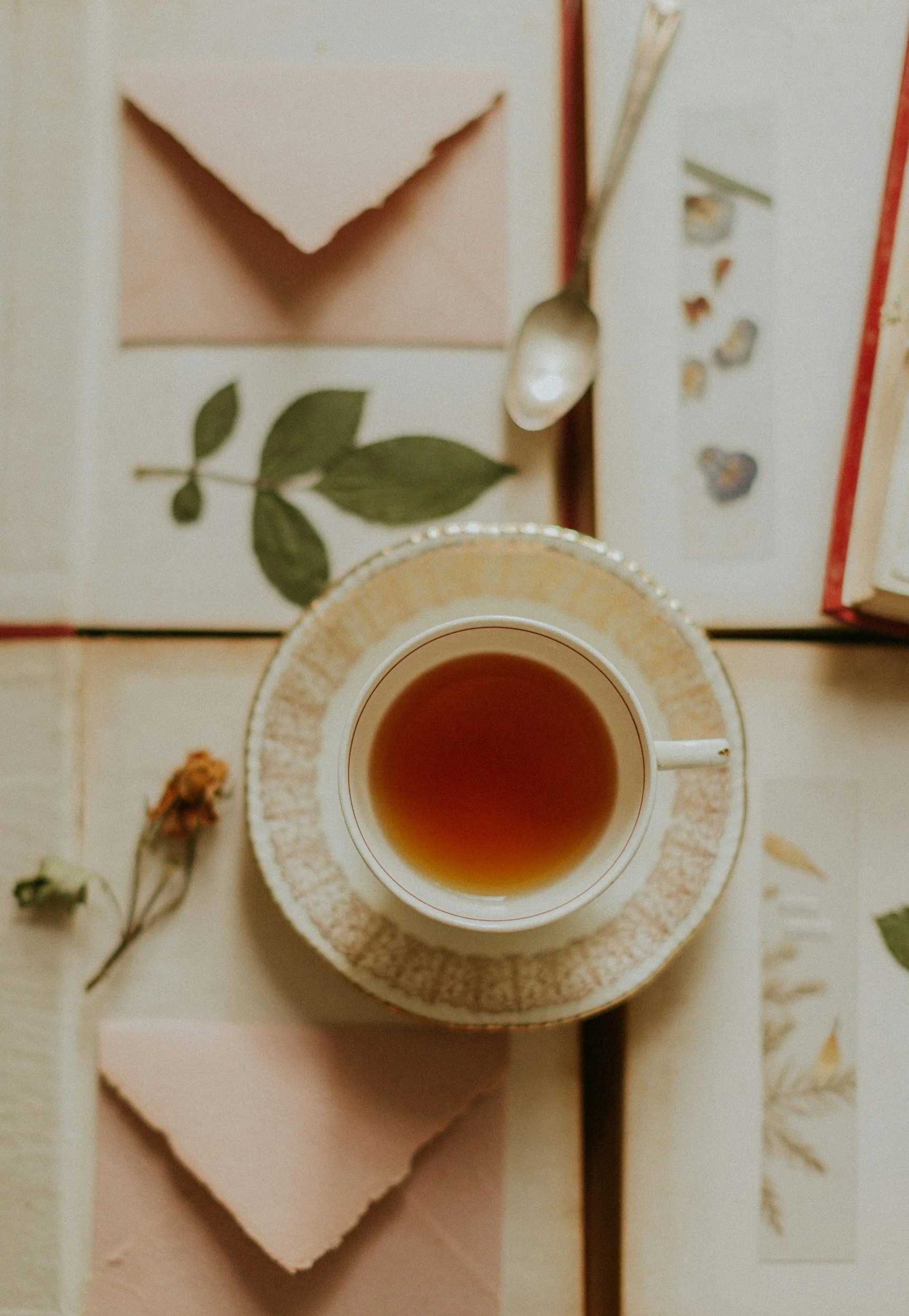 A cup of tea on a saucer, surrounded by dried flowers, a spoon resting on a napkin, and a few scattered tea leaves on a textured surface with floral patterns.