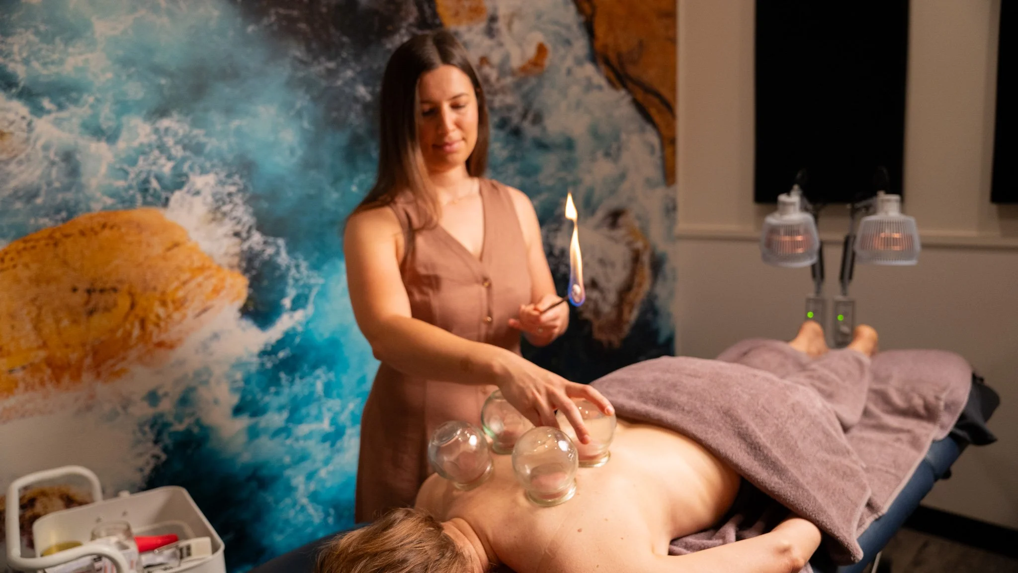 cupping therapy wollongong - A woman in a brown dress performs a cupping therapy session on a person lying face down on a massage table, with glass cups placed on their back, during a treatment in a room with ocean-themed wall art.