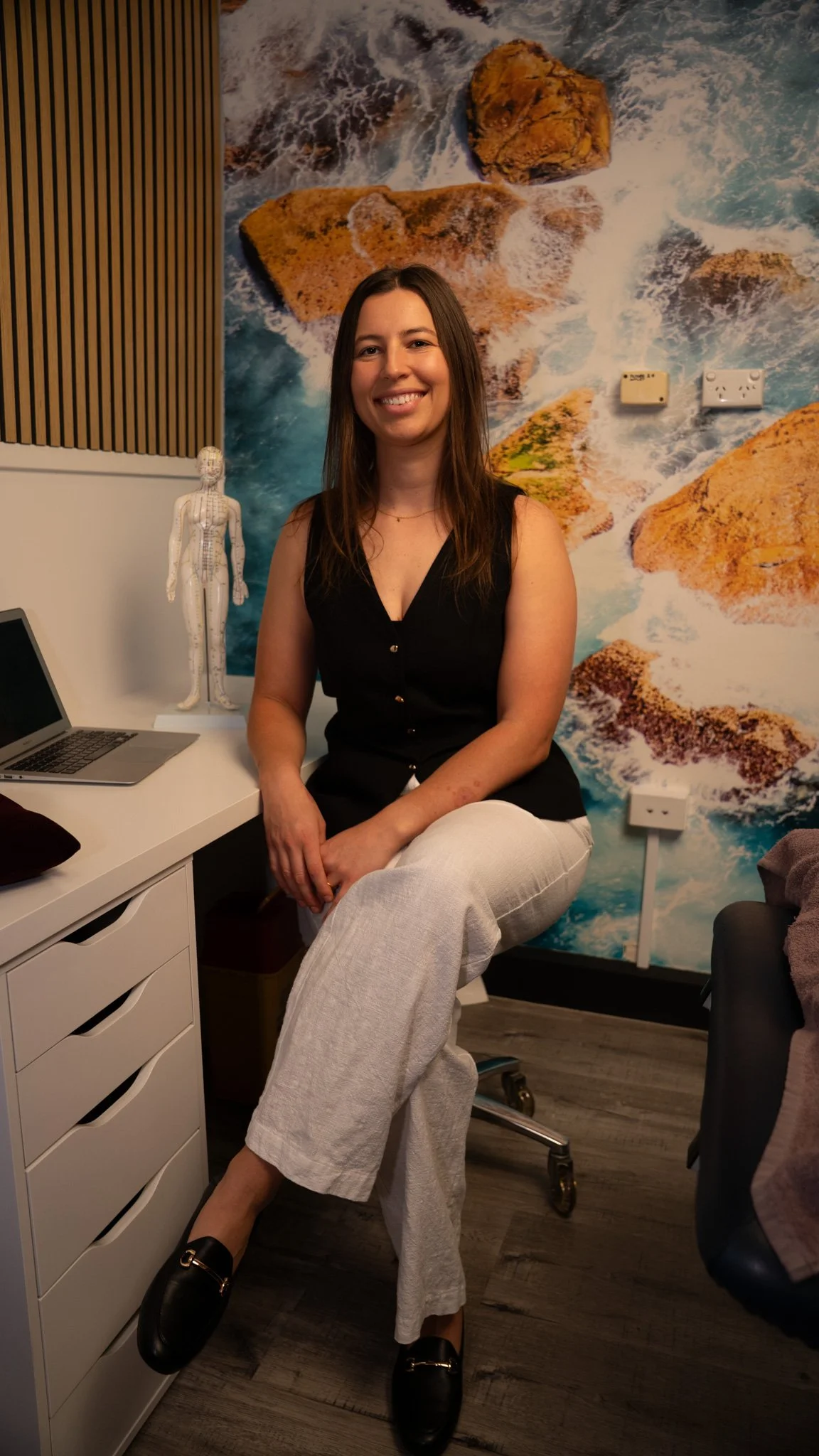 acupuncture near me wollongong - A woman sitting on a chair in an office, smiling, wearing a black top, white pants, and black shoes.