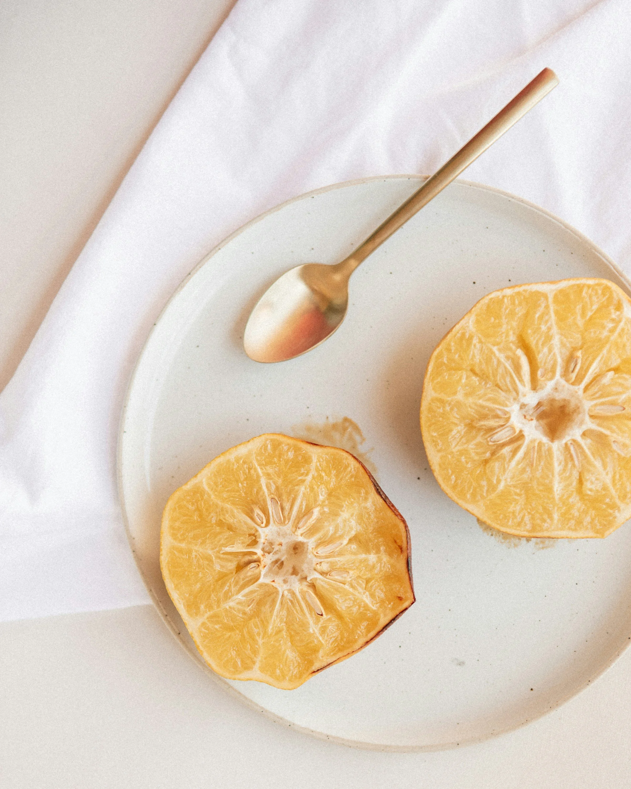 Close-up of halved oranges on a white plate with a gold spoon, on a light-colored fabric background.