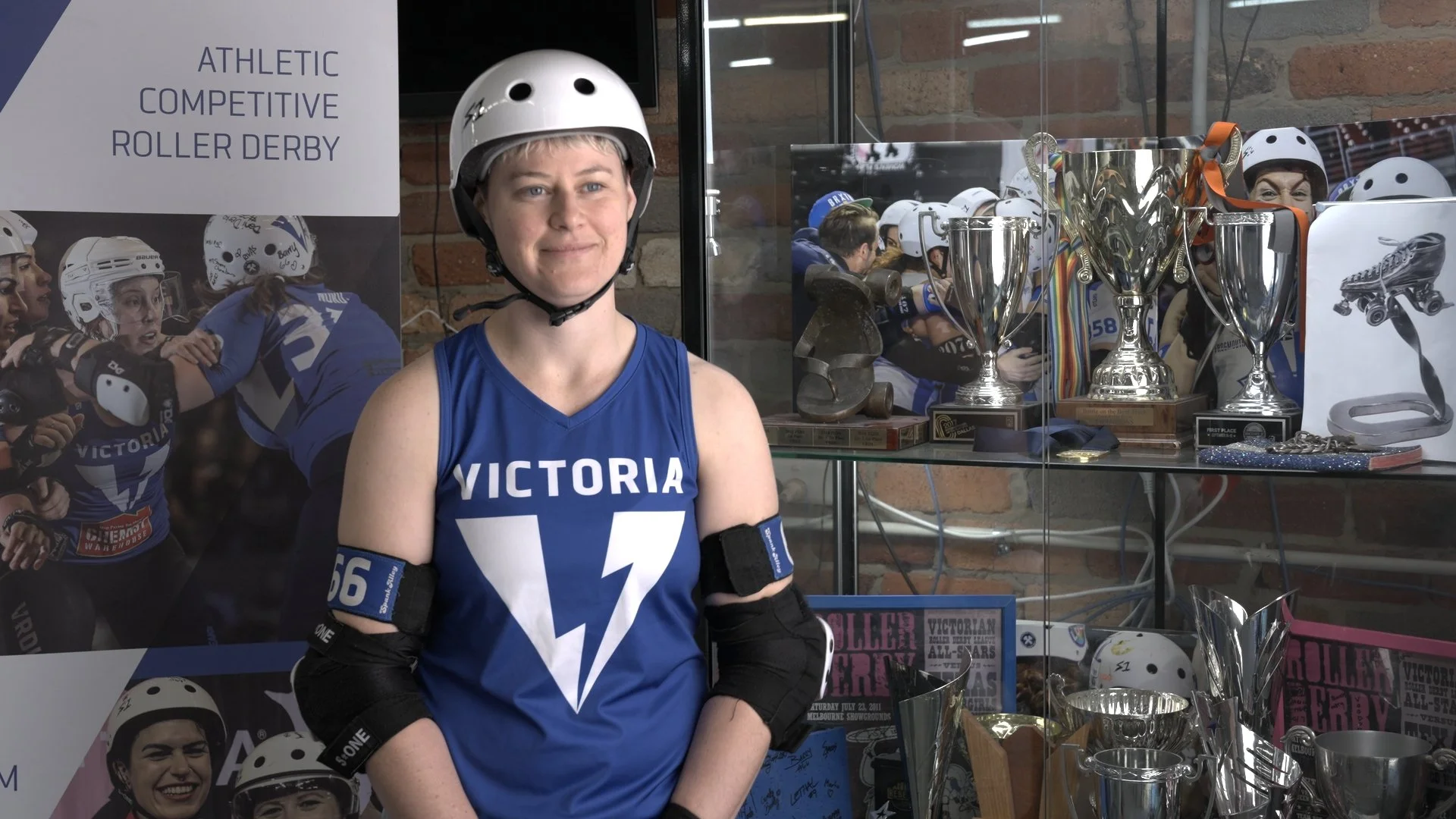 Woman wearing a blue Victoria roller derby jersey and helmet, standing in front of a display case with trophies and roller derby memorabilia at a roller derby event.