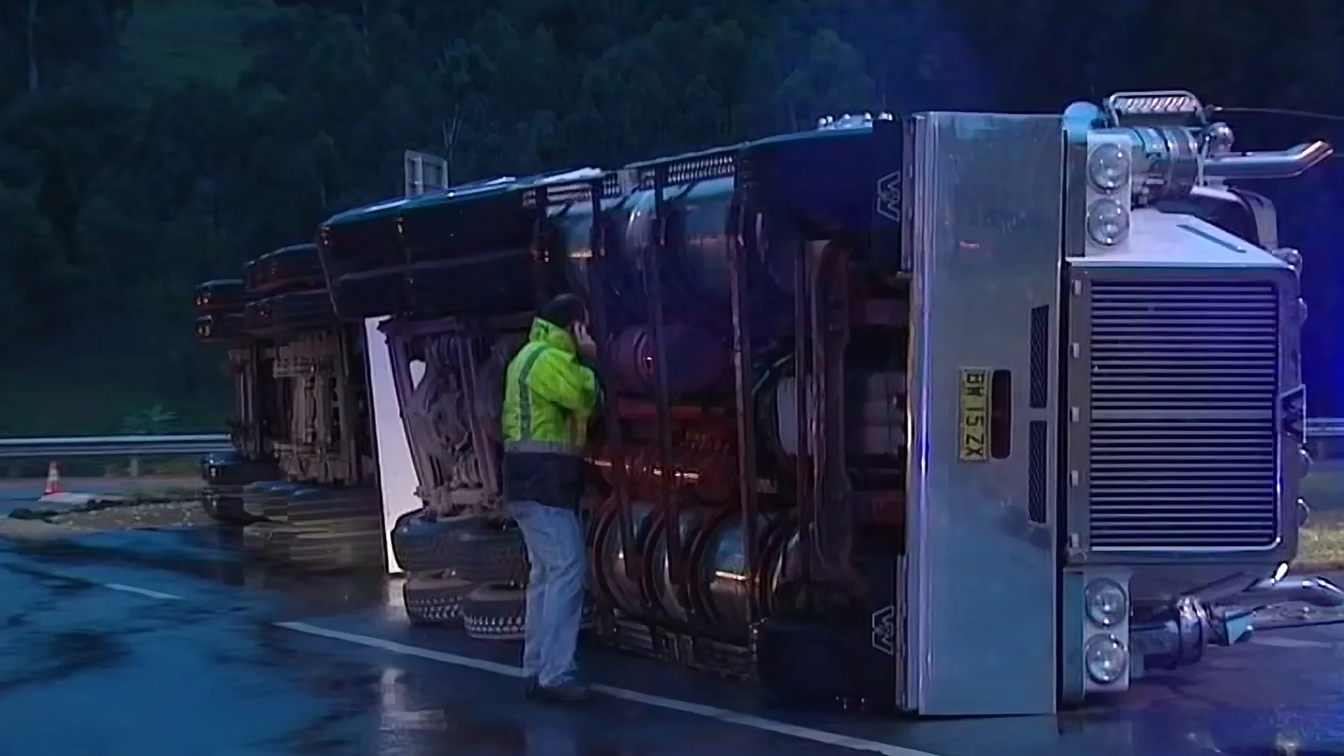 A large truck is flipped on its side on a wet road with a person in a yellow safety vest inspecting the vehicle.