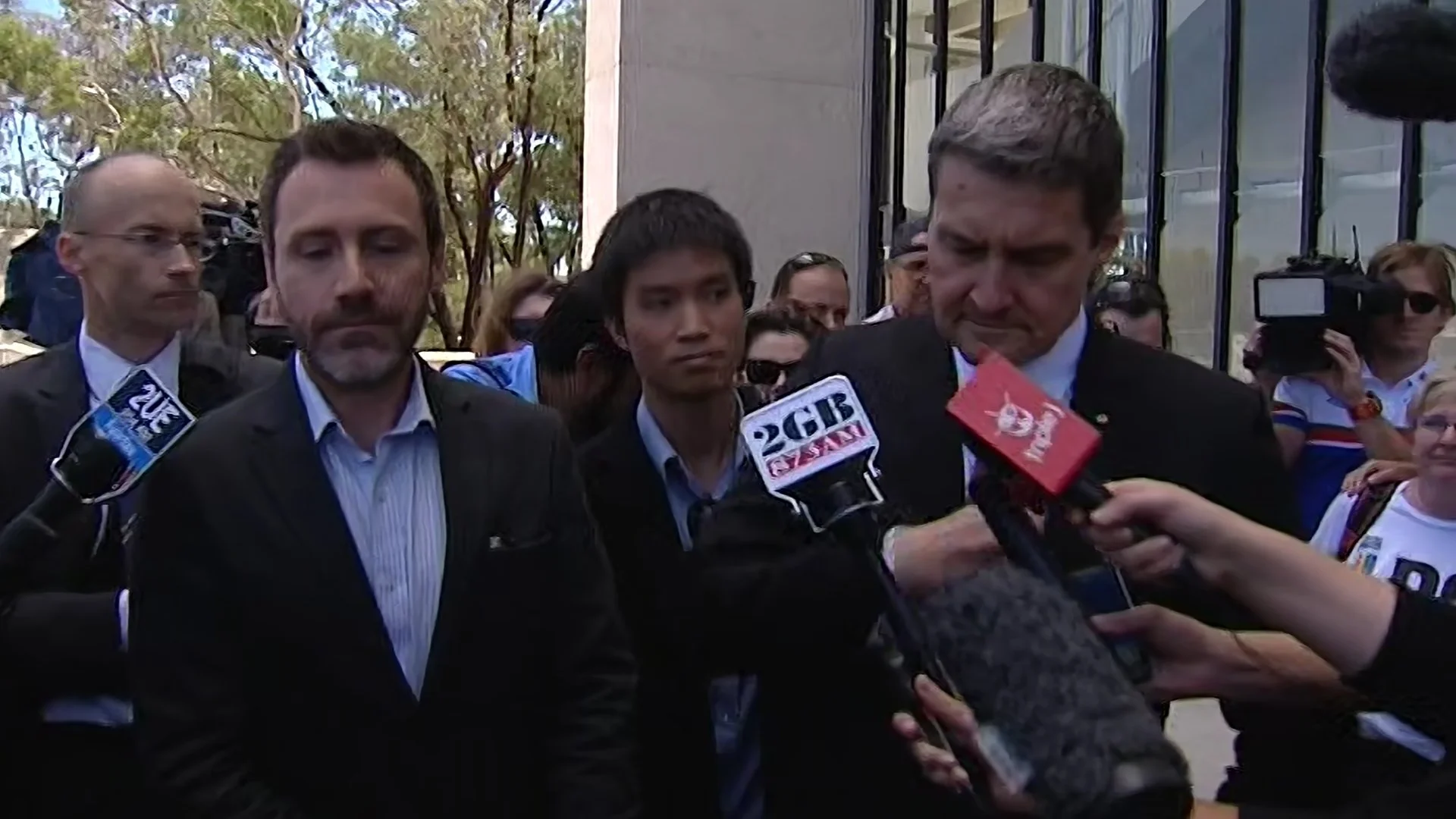 Group of three men in suits being interviewed by multiple reporters with microphones, outdoors with trees and a modern building in the background.