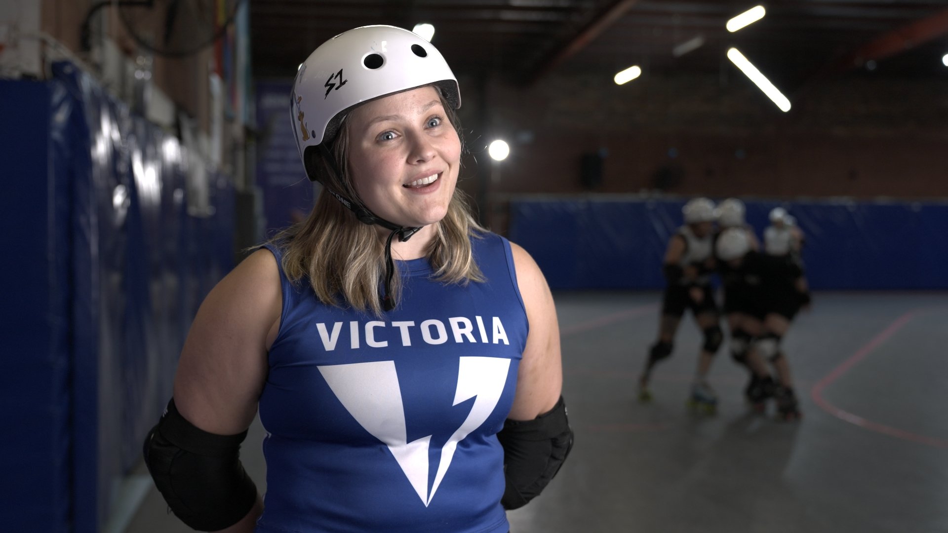 Female roller derby athlete wearing a white helmet and a blue jersey with 'Victoria' and a thunderbolt logo, standing in an indoor roller derby rink with other players in the background.
