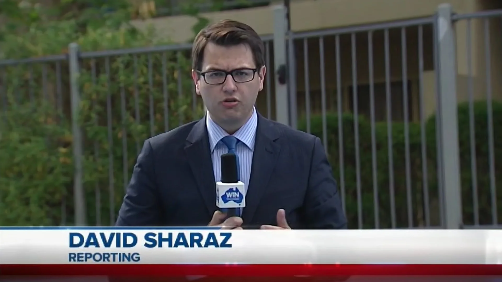 News reporter named David Sharaz reporting outdoors with a microphone in hand, wearing glasses and a dark suit, standing in front of a fenced area with green foliage.