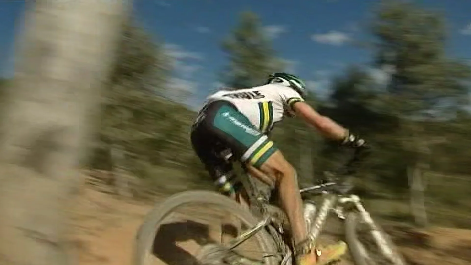 A mountain biker in a white and teal jersey riding a bike on a dirt trail, surrounded by trees with a blue sky overhead.