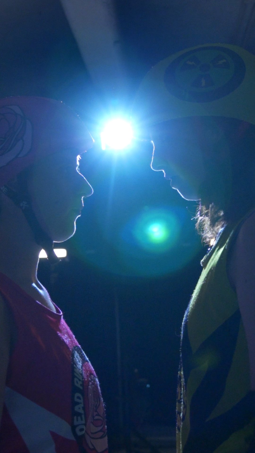 Two female ice climbers face each other in profile against a bright light, wearing helmets and gear.