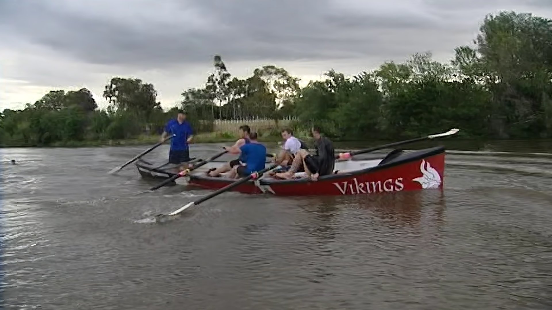 A group of six people in a red Viking-themed rowboat on a river, with a woman taking a selfie and others sitting or standing, during cloudy weather.