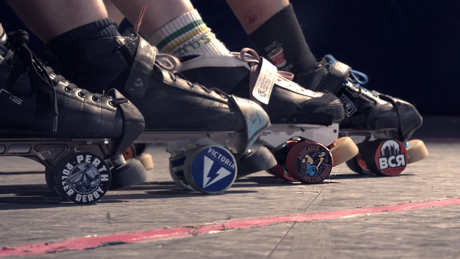 Close-up of four roller skates with colorful stickers on the wheels, positioned on a concrete surface with a pink line, outside.
