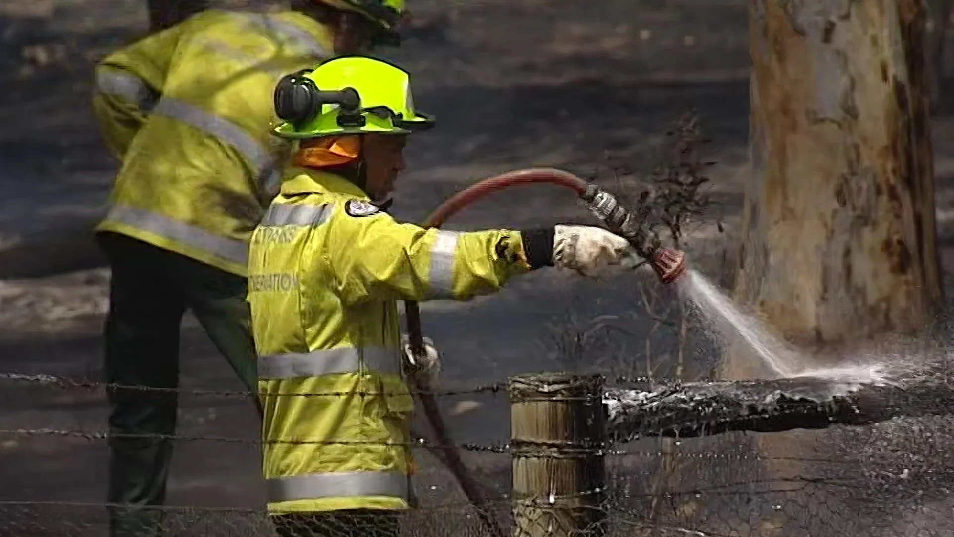 Firefighter in yellow protective gear and helmet using a hose to spray water on a burnt area.