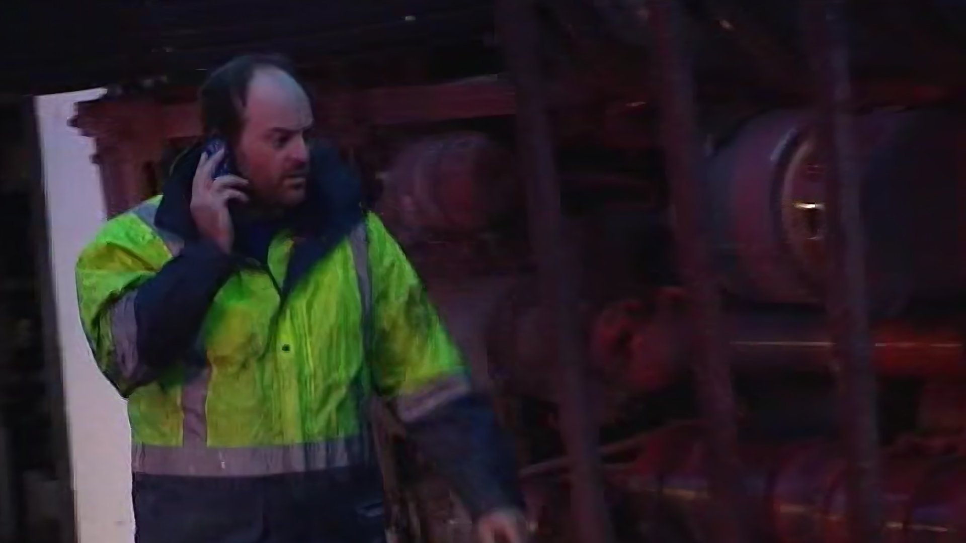 A man in a bright yellow-green high visibility jacket speaking on a cell phone in a dark environment with blurred machinery in the background.
