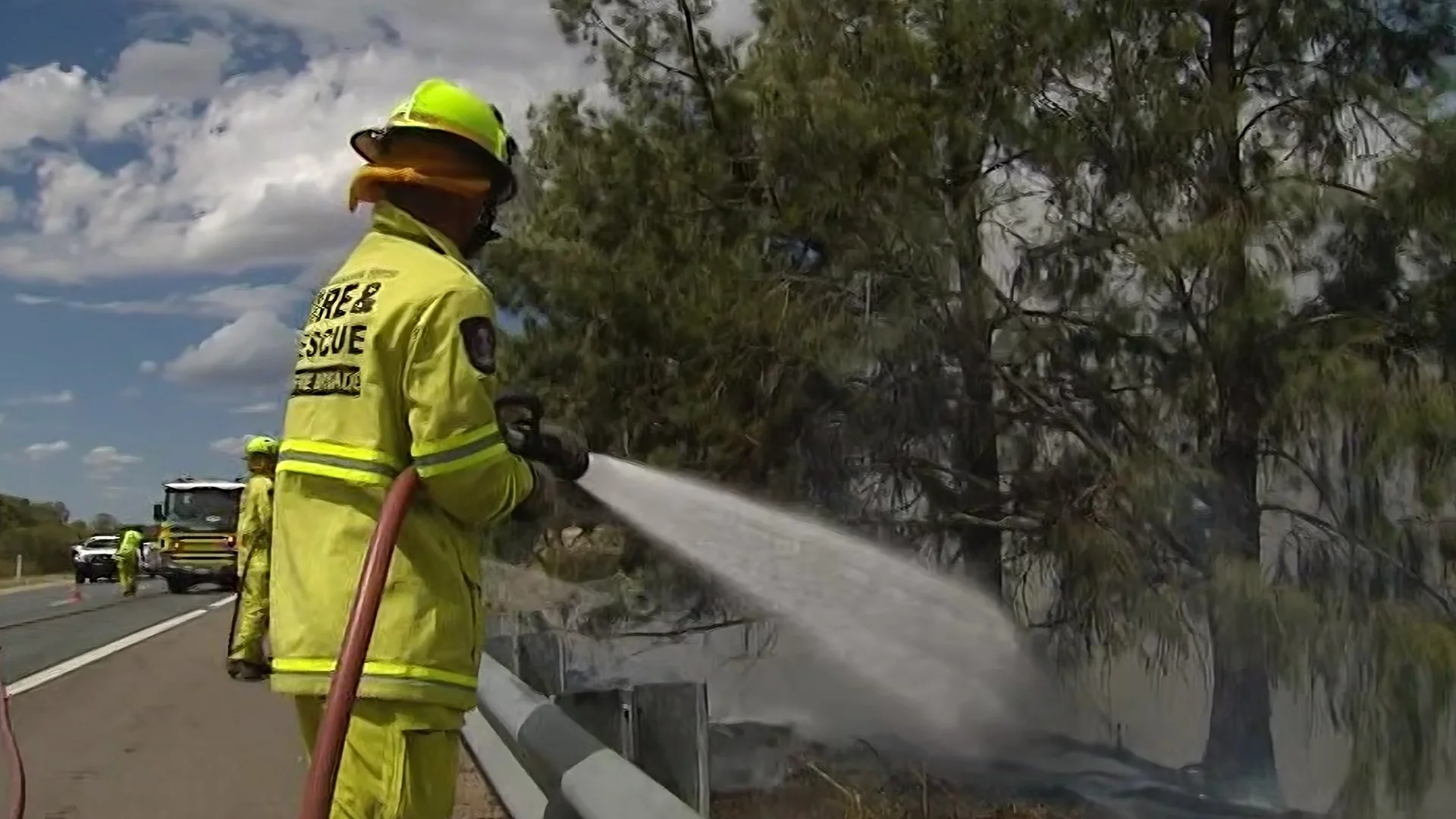 Firefighters in yellow protective gear and helmets are using a hose to spray water on a large fallen tree that is partially blocking a roadway, with emergency vehicles and personnel in the background under a partly cloudy sky.