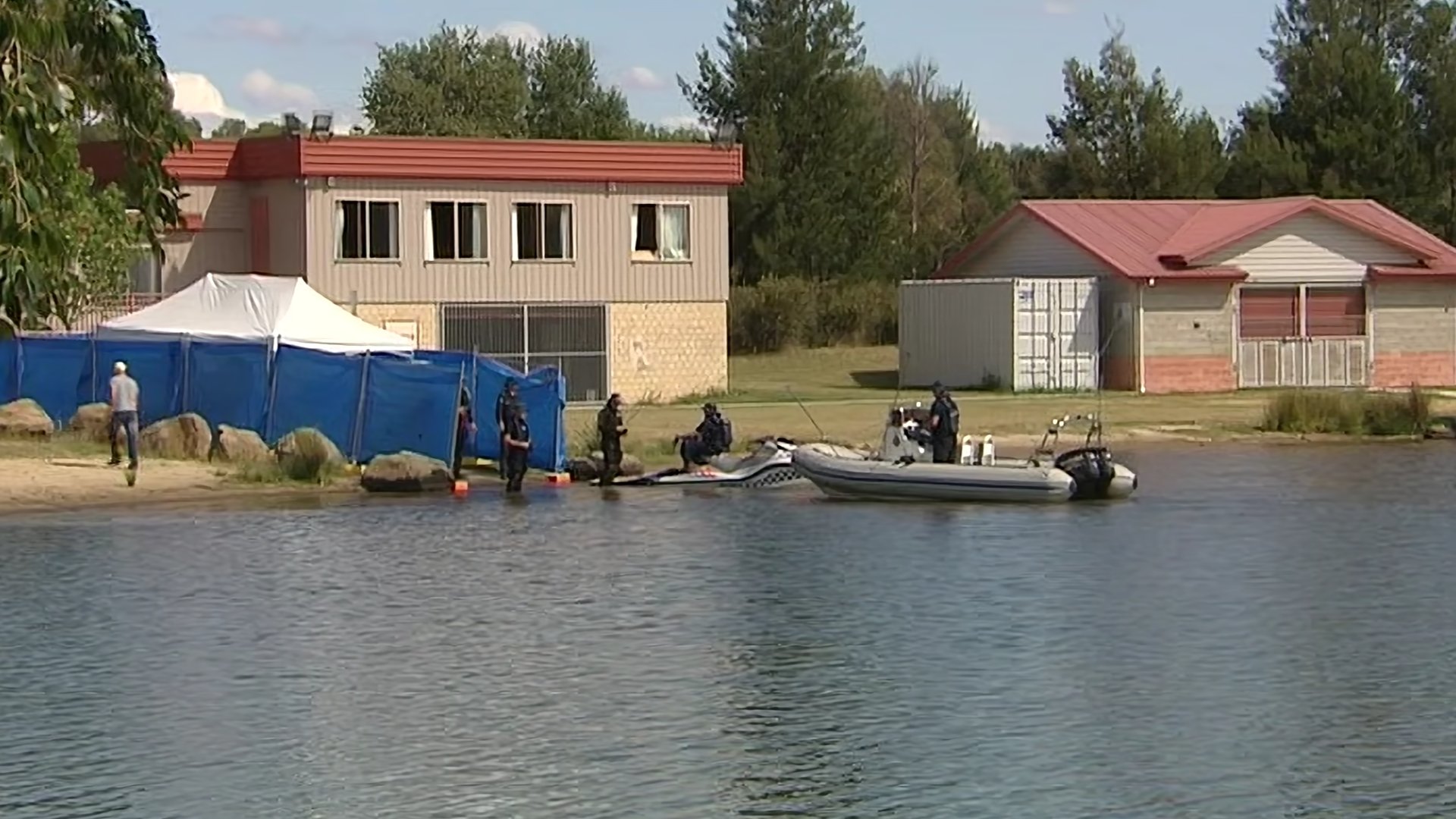 People in water near a blue fence, with houses and trees in the background, on a sunny day.