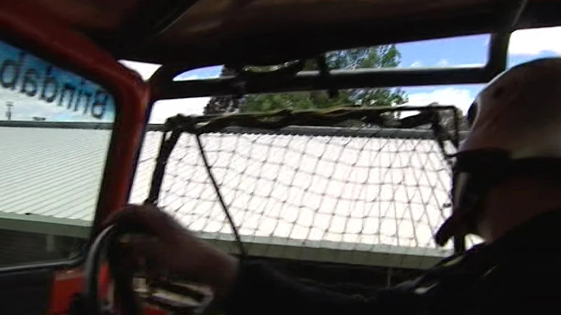 Interior view of a race car or go-kart with a mesh safety net in front of the windshield. The windshield has a sticker on the left side, and there is a person wearing a helmet or hat on the right side. The background shows trees and a blue sky.