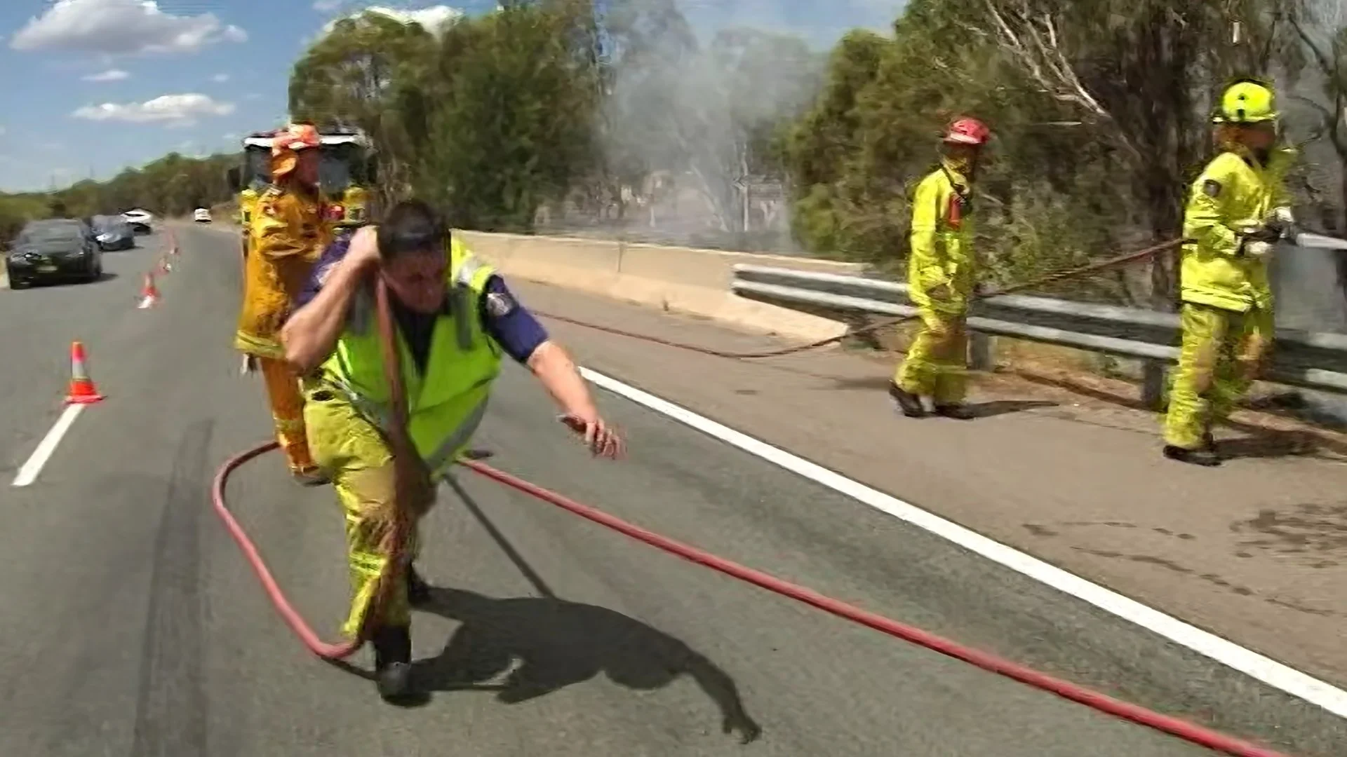 Firefighters in yellow protective gear and helmets working on a highway with smoke and damaged trees in the background.