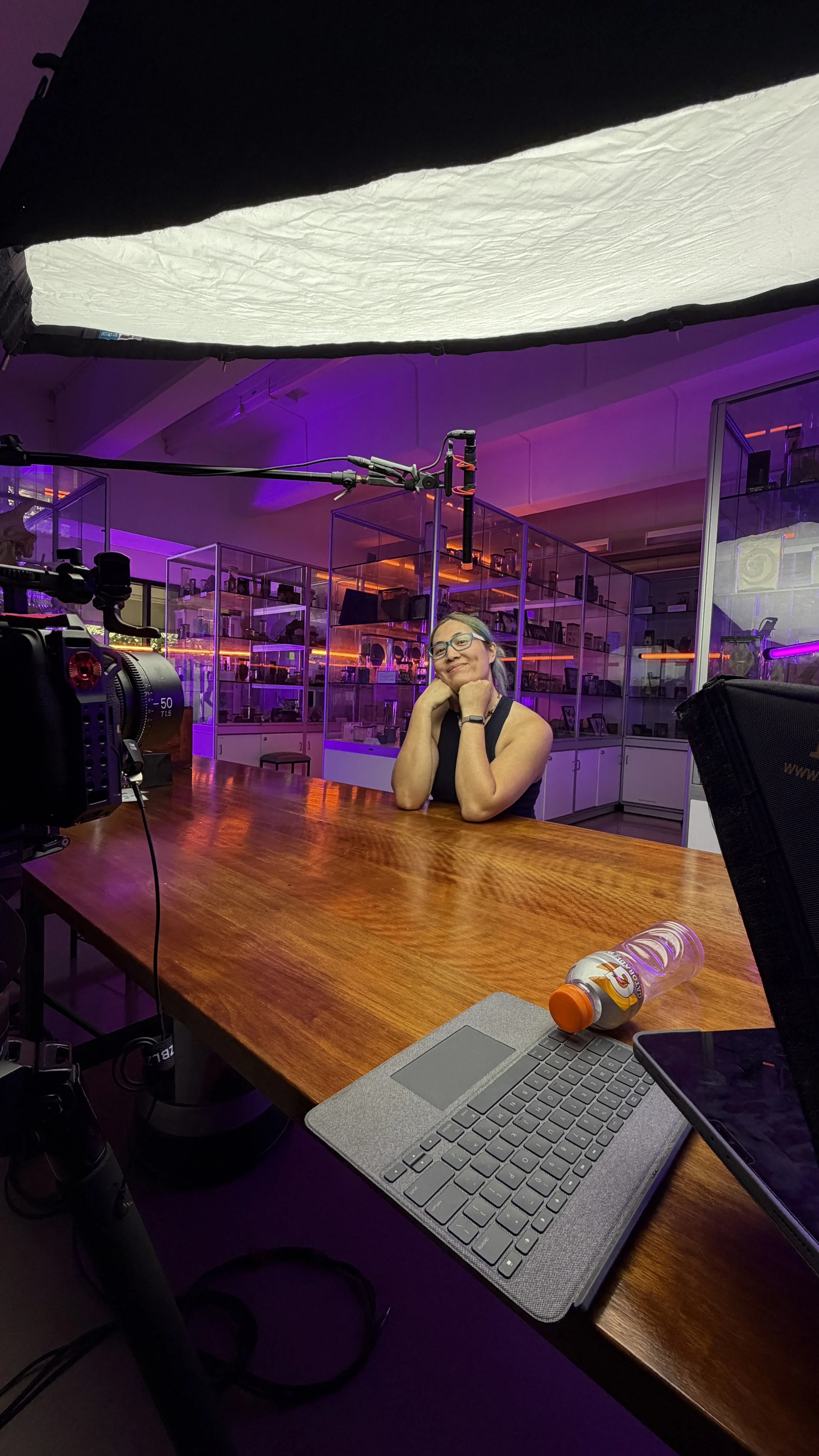 A woman sitting at a wooden table in a modern, brightly lit room with purple and orange lighting, behind glass display cabinets. There is a camera, a laptop, and a water bottle on the table.