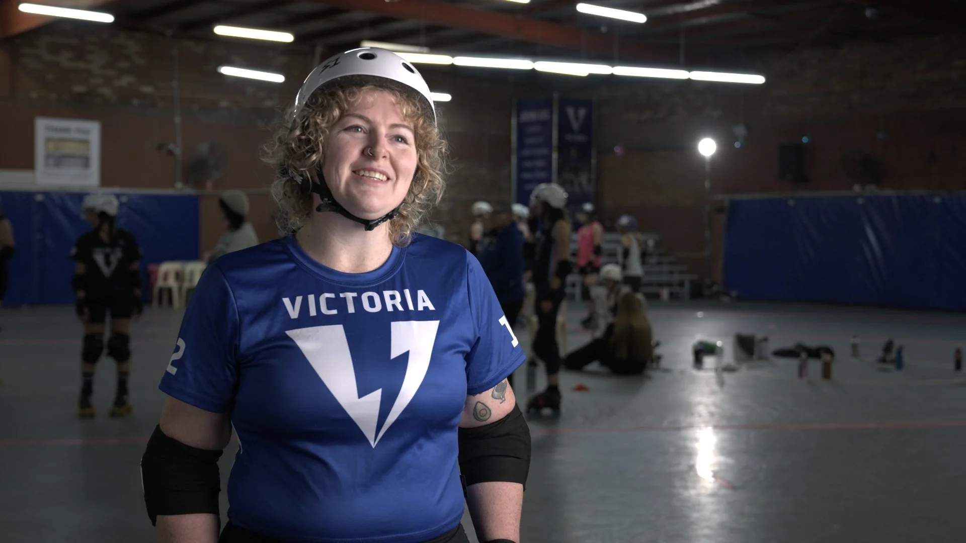 A young woman with curly blonde hair smiling in a roller derby rink, wearing a blue Victoria jersey and a white helmet, with other skaters and equipment in the background.