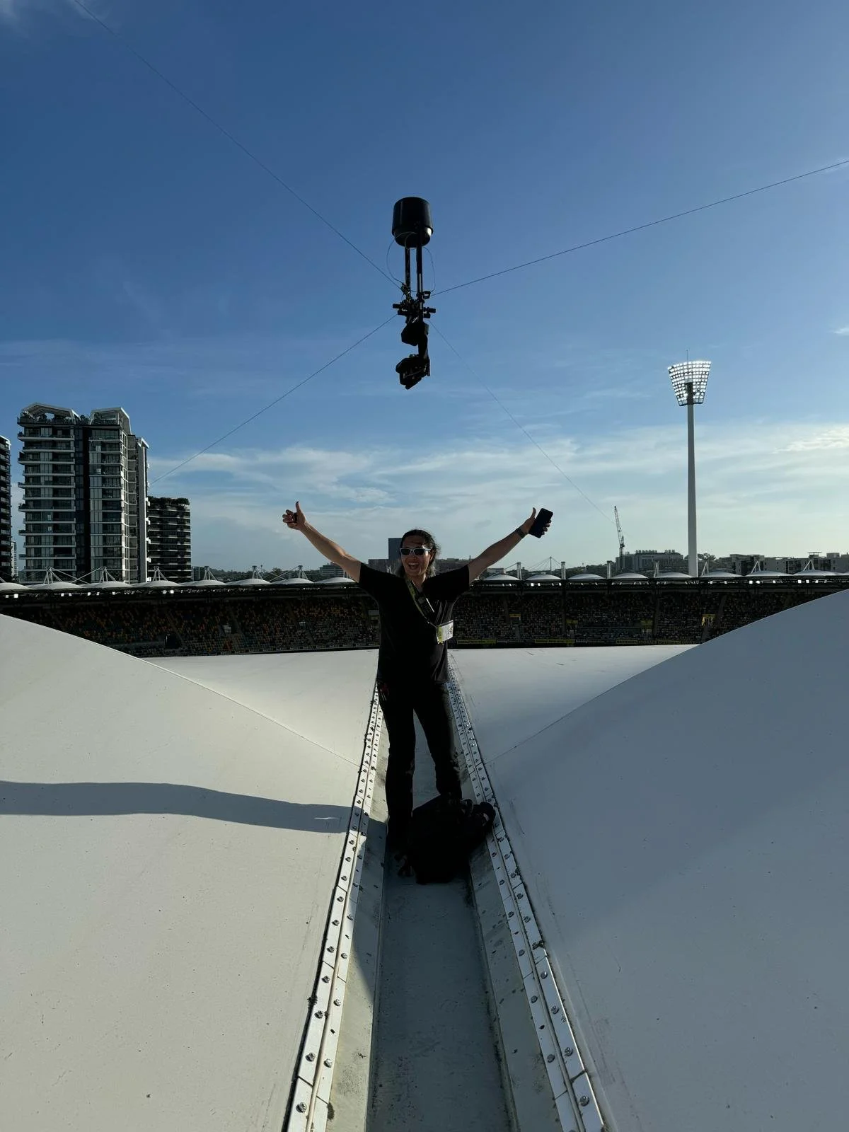 A person standing on top of a large, white, curved structure at a stadium, smiling and raising their arms in celebration. The sky is blue with a few clouds, and there are high-rise buildings and a tall floodlight tower in the background.