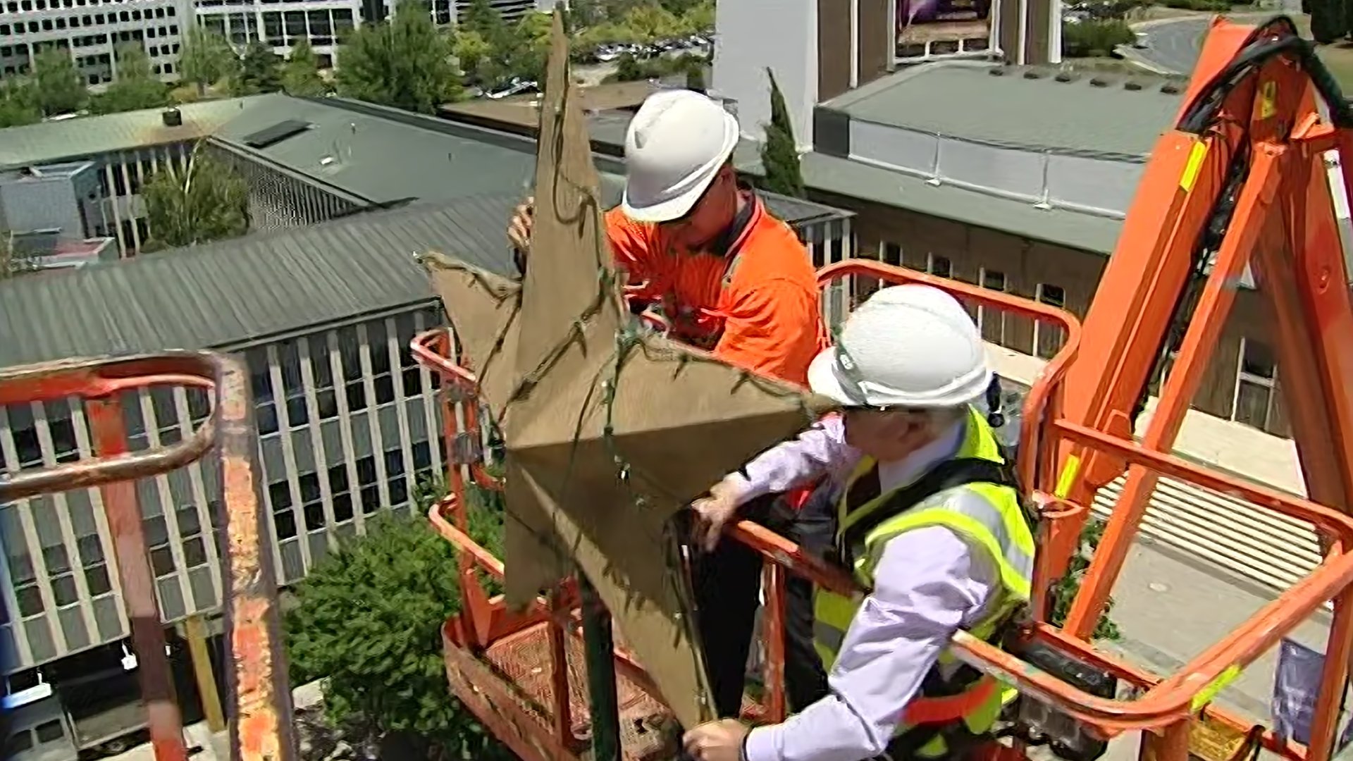 Workers wearing safety helmets and vests on an orange lift platform trimming a tree.