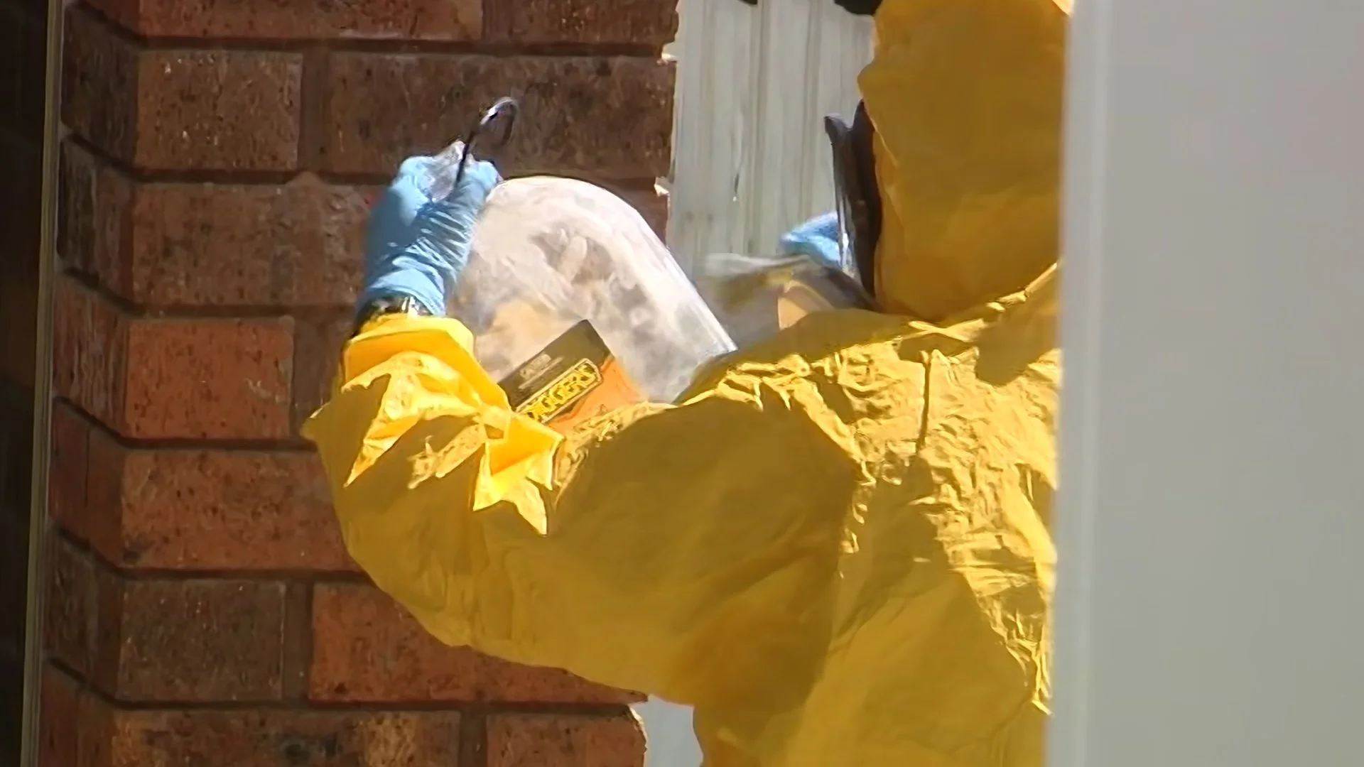 A person's hands wearing blue gloves and a yellow raincoat are applying a layer of mortar or cement to red brick wall with a trowel.