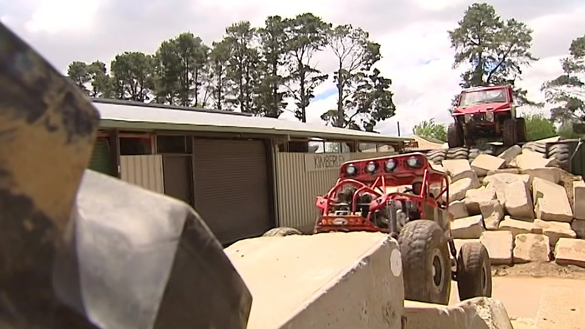 Red off-road vehicles stacked on rocks and a driveway with trees in the background.