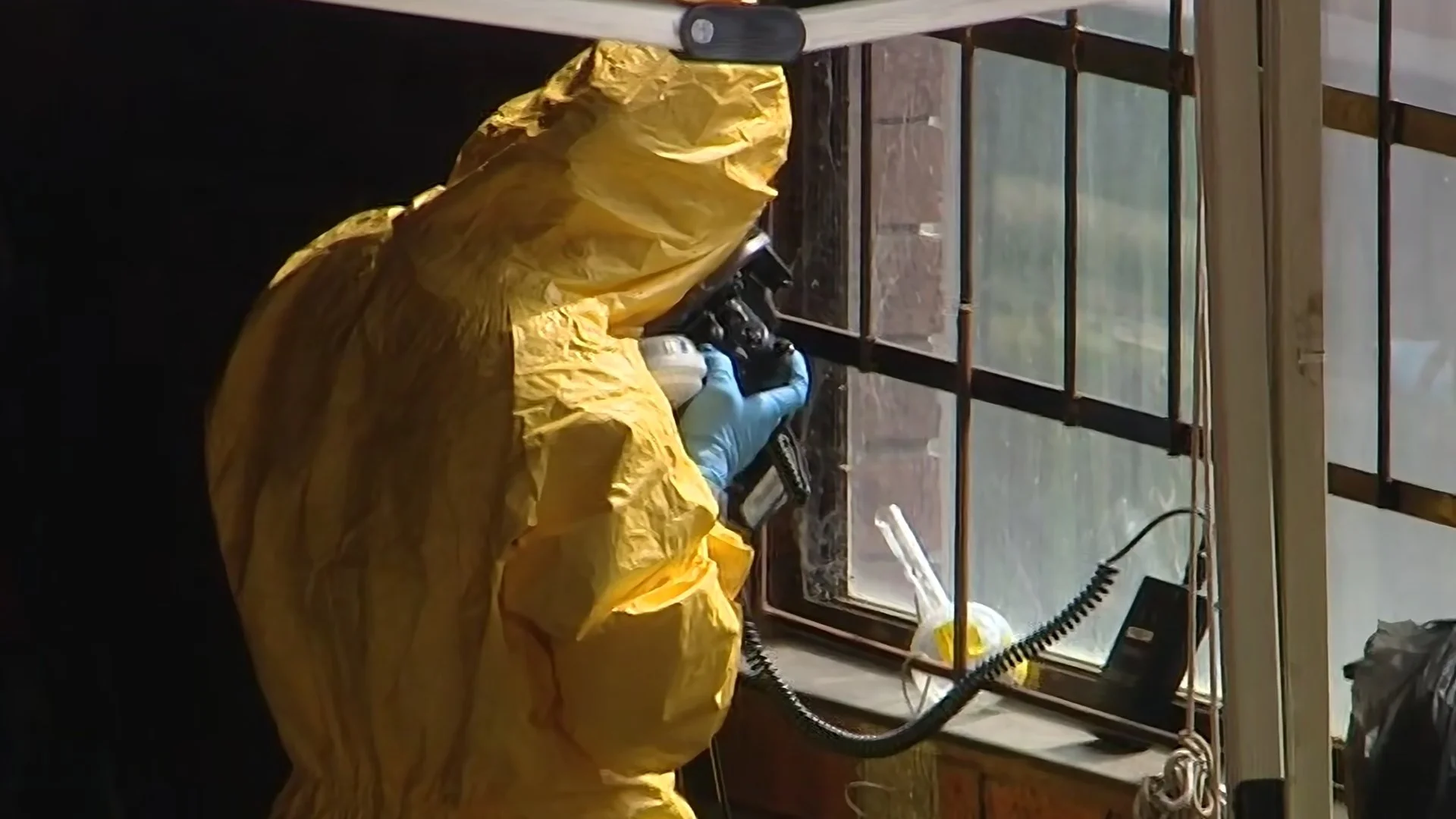 Person wearing a yellow protective suit and blue gloves inspecting or repairing equipment near a window with sunlight coming through.
