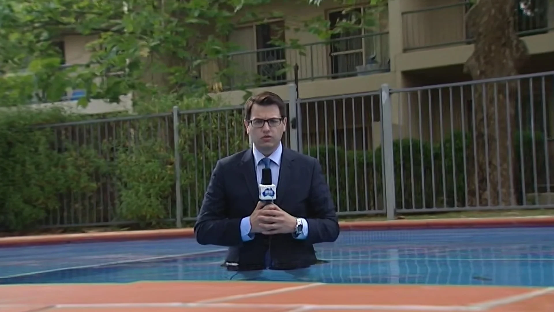 Male news reporter in a suit and glasses holding a microphone, standing outdoors behind a swimming pool with a metal fence and green trees in the background.